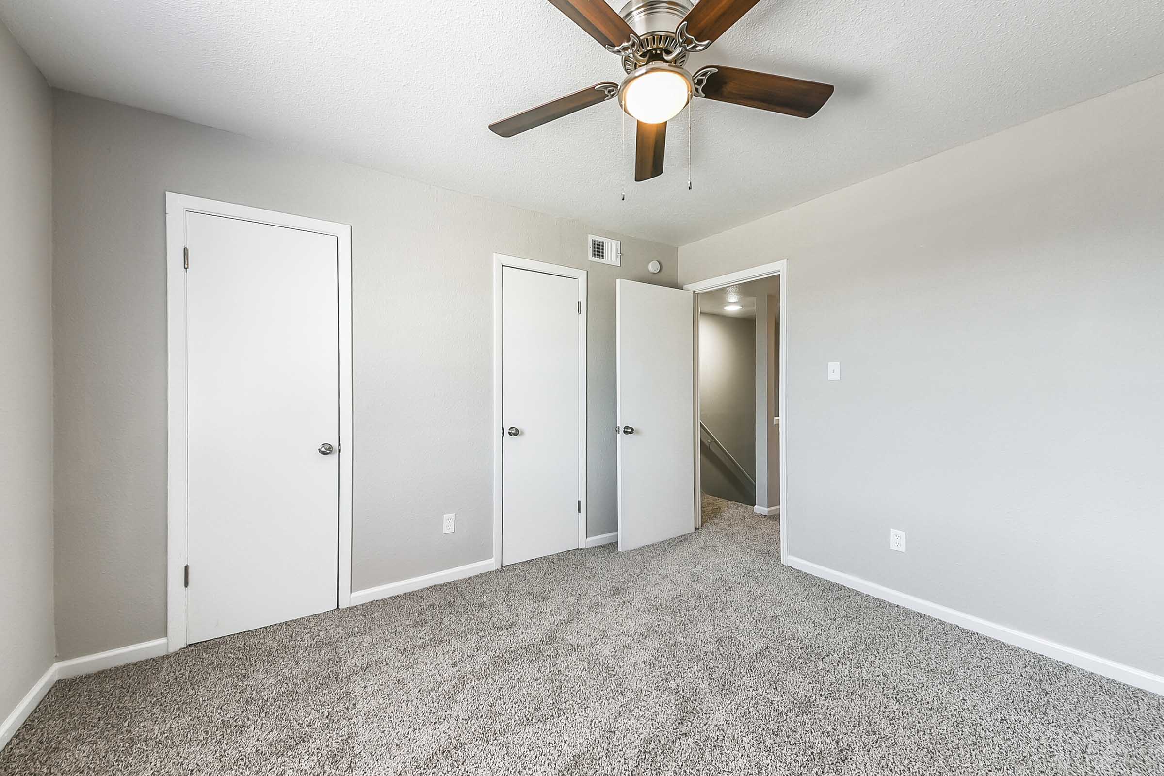 A well-lit bedroom featuring light gray walls and plush carpet. The room includes a ceiling fan with wooden blades, two white doors leading to closets, and a third door that appears to lead to another room or hallway. The overall space is simple and modern, with neutral tones.