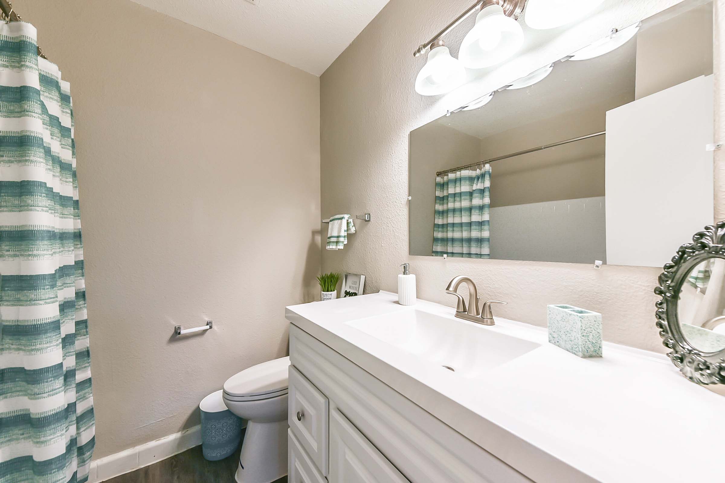 A modern bathroom featuring a white vanity with a sink, a large mirror, and a colorful striped shower curtain. The space includes a toilet, hand towel, and decorative items like a plant and a light blue storage box, with soft lighting from above.