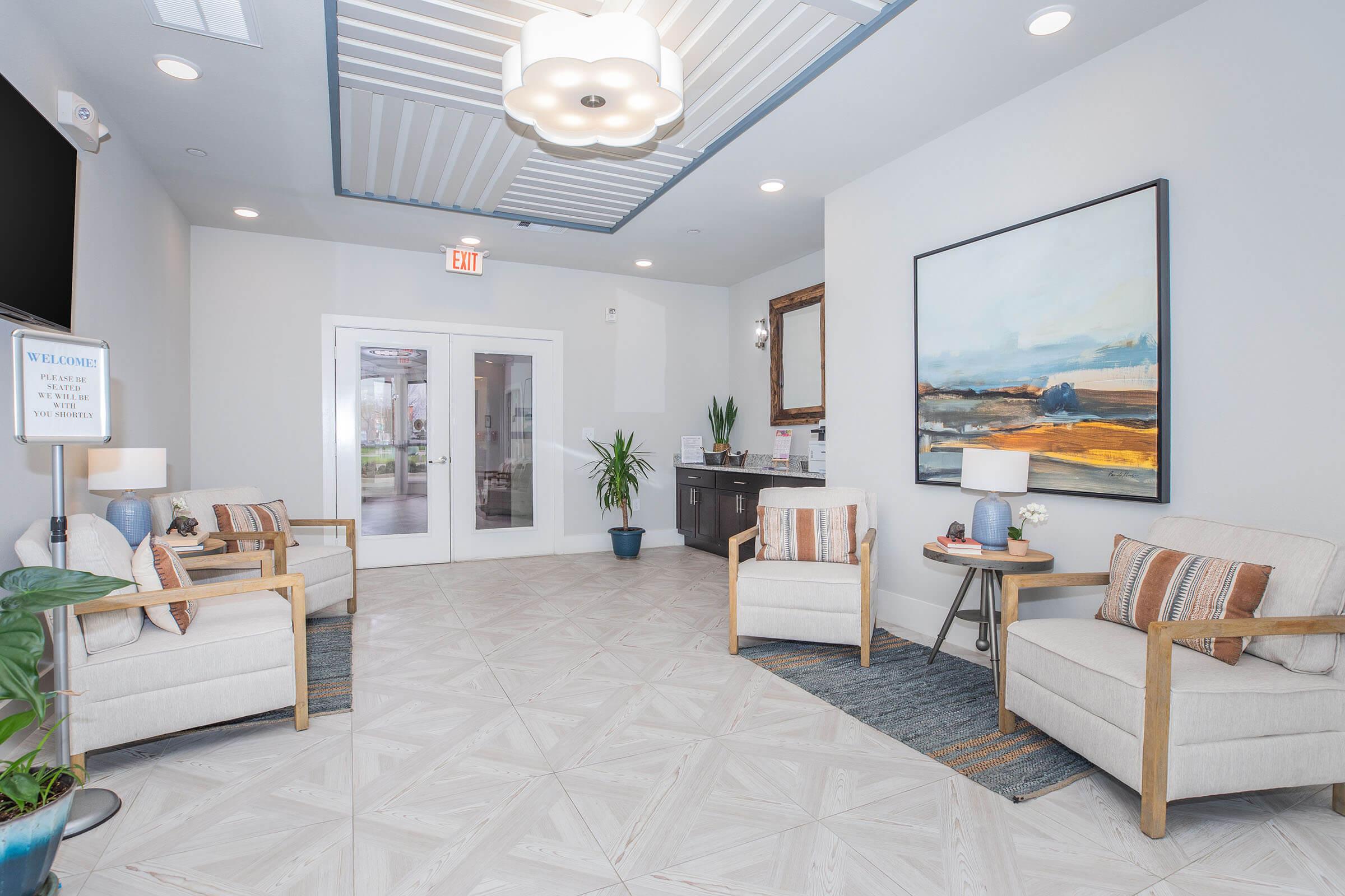 A modern reception area featuring two beige armchairs with orange throw pillows, a large abstract painting on the wall, a small table with lamps, and potted plants. The space is well-lit with a ceiling light and has light-colored flooring with a patterned design. There are glass doors leading to another room.