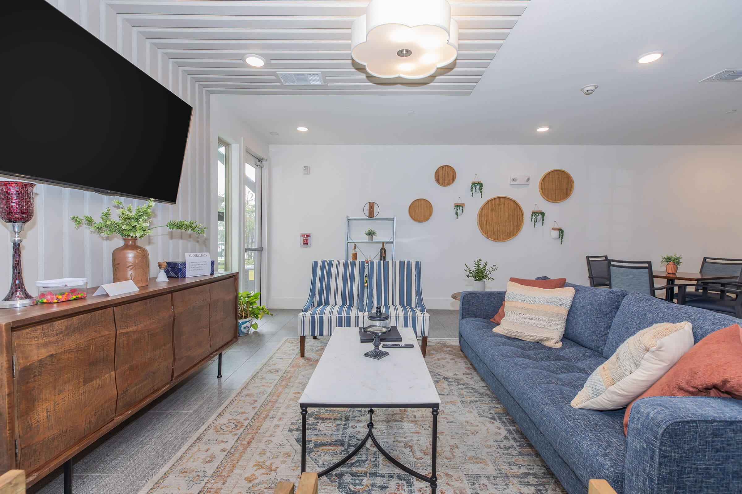 A modern living room with a blue sofa and striped chairs, a coffee table with a decorative vase, and a wooden sideboard. The walls feature round wooden accents, and there is a large television in the corner. Natural light fills the space, enhancing the cozy atmosphere.