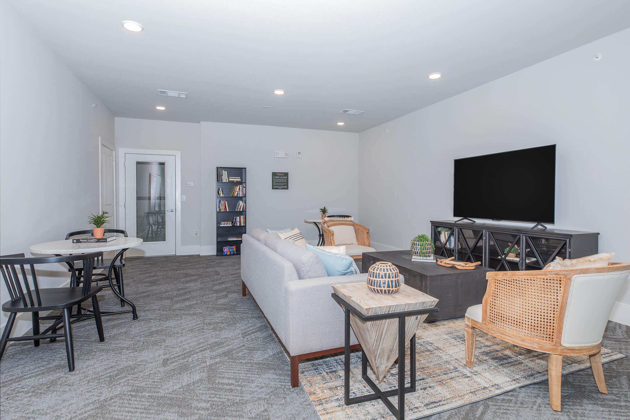 Modern living room featuring a light gray sofa with decorative pillows, a coffee table, and a stylish TV stand with a large TV. A small dining area with a round table and two chairs is visible in the corner. The walls are painted in neutral tones, and there are bookshelves on the wall, adding a cozy touch to the space.