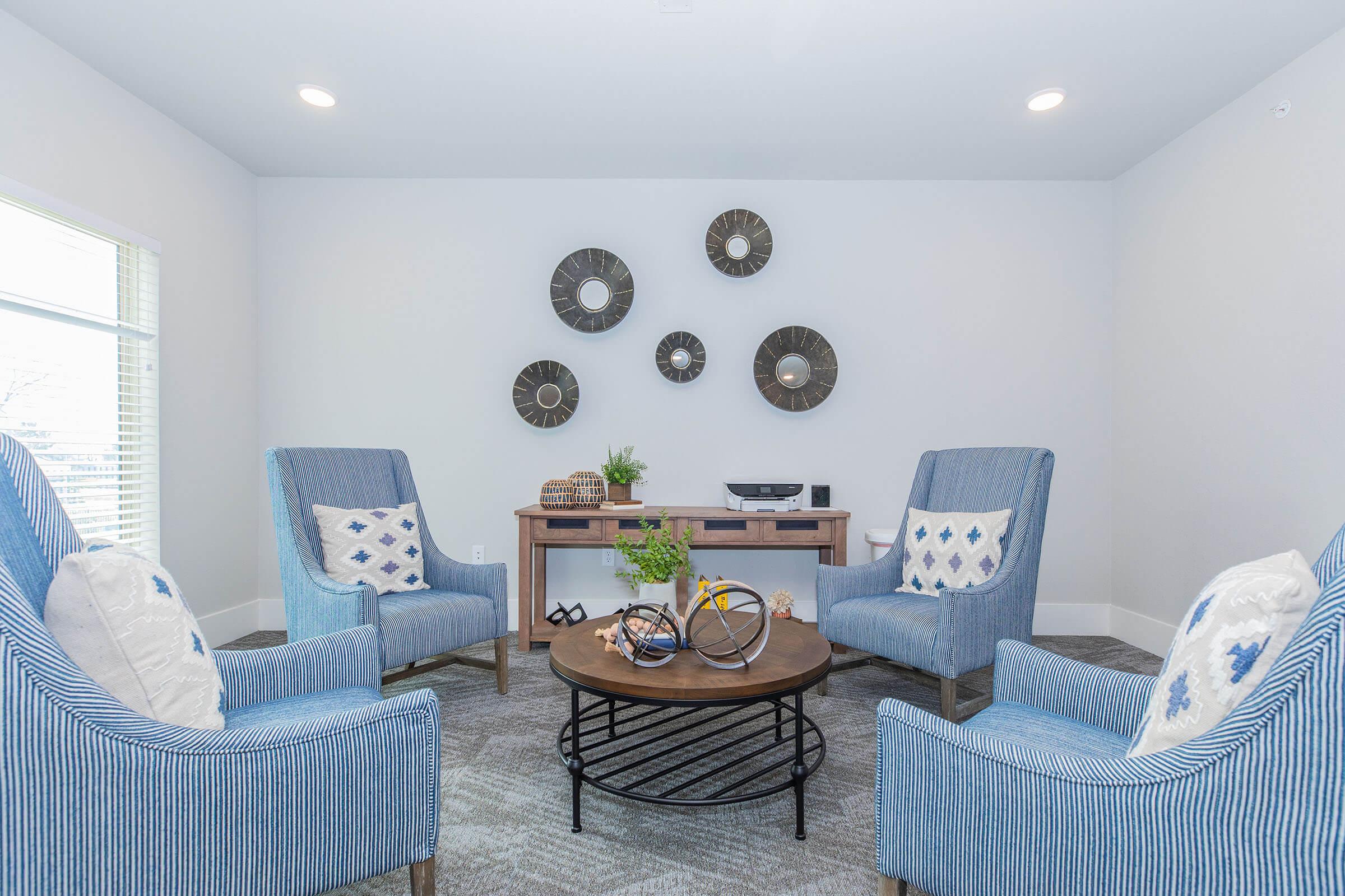 A cozy living room featuring two blue striped armchairs with decorative pillows, a round coffee table in the center, and a wooden console table. Above the console, there are three circular wall mirrors. Decorative items like a plant and a bicycle sculpture are arranged on the table, set against a light gray wall and carpet.