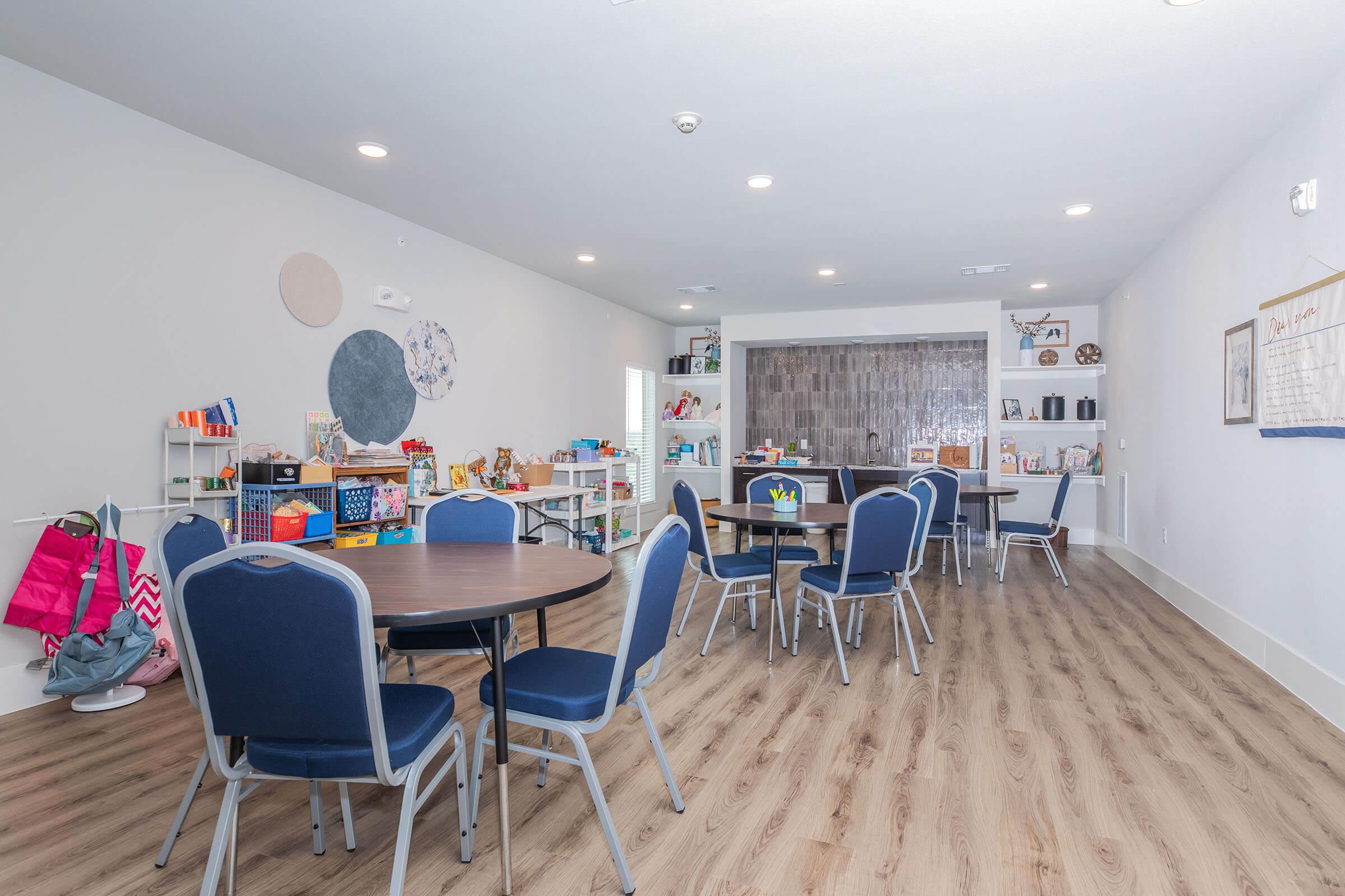 A bright, spacious room with several round tables and chairs arranged for social or educational activities. The walls are light-colored, and there's a kitchenette area in the background. Various shelves hold toys and educational materials, creating a welcoming environment for gatherings or learning.