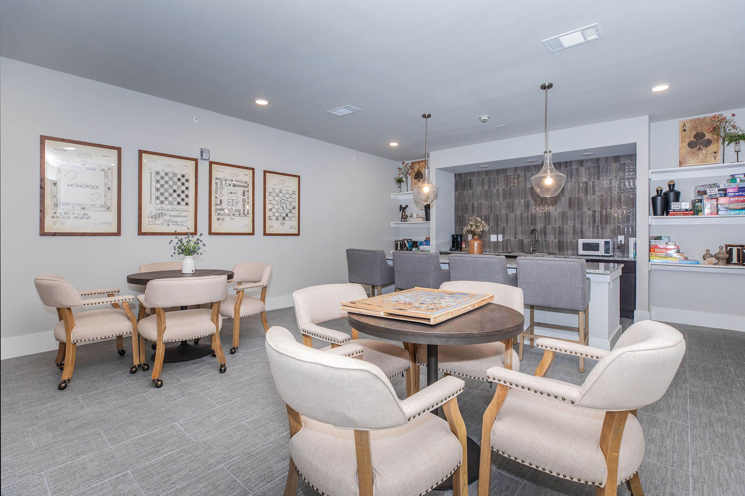 A modern communal lounge area featuring two round tables with four chairs each, a kitchenette in the background, and framed artwork on the walls. Soft lighting from pendant lamps adds warmth to the space, which is decorated with shelves holding books and games. The flooring is light gray and the walls are neutral.
