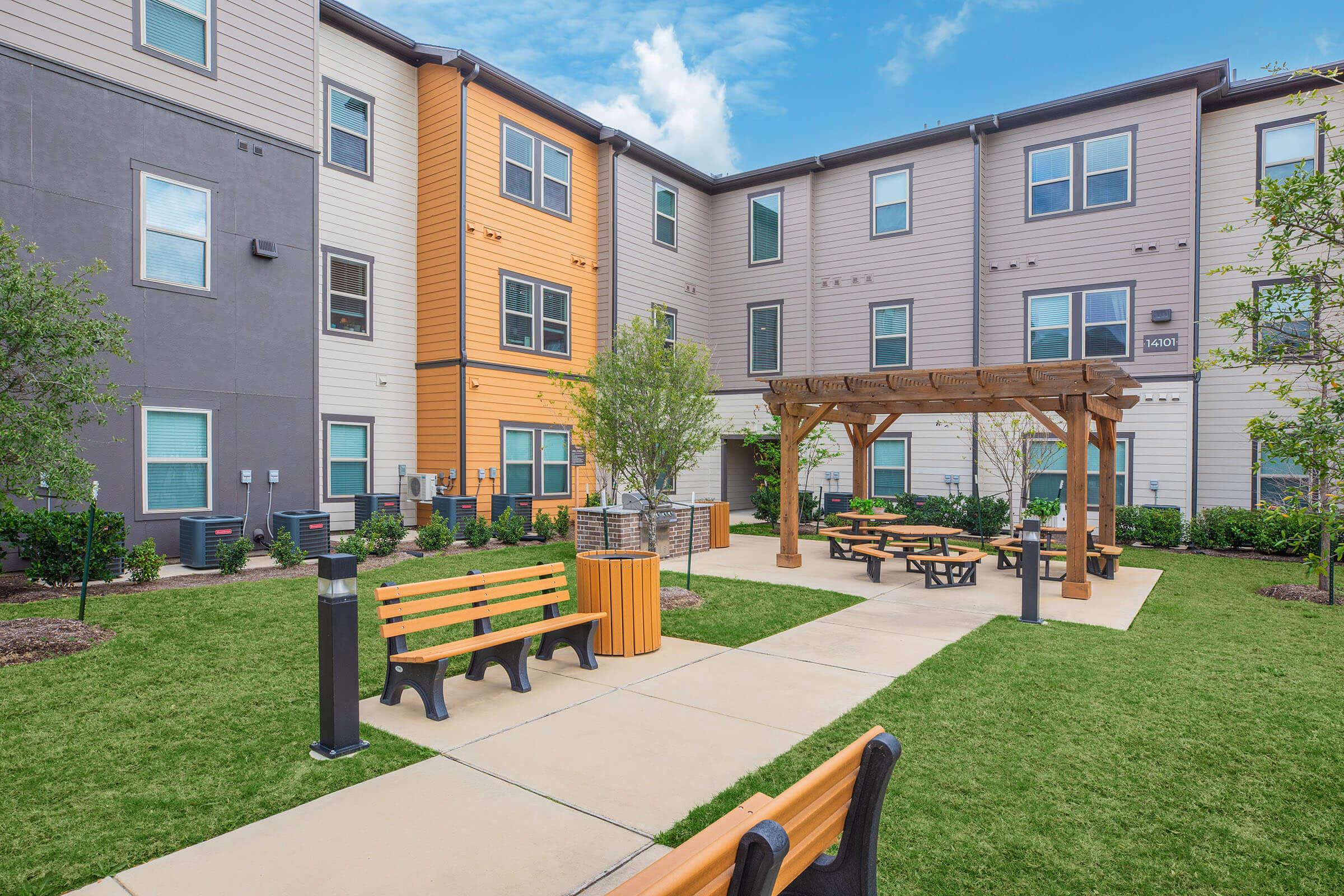 A landscaped courtyard with green grass, benches, picnic tables, and a wooden pergola. Surrounding the area are modern multi-story residential buildings in various colors, creating a welcoming community space for residents.