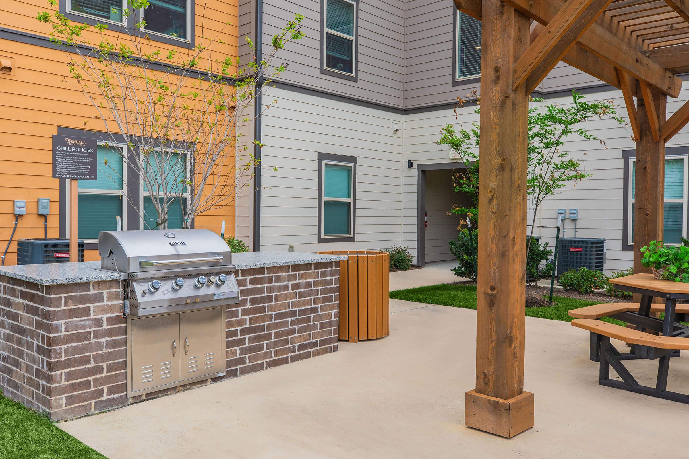 Outdoor grilling area with a stainless steel barbecue grill situated on a stone and concrete platform. Nearby, there are wooden picnic tables under a pergola, with green grass and shrubs adding greenery to the space. The area is designed for relaxation and outdoor dining.