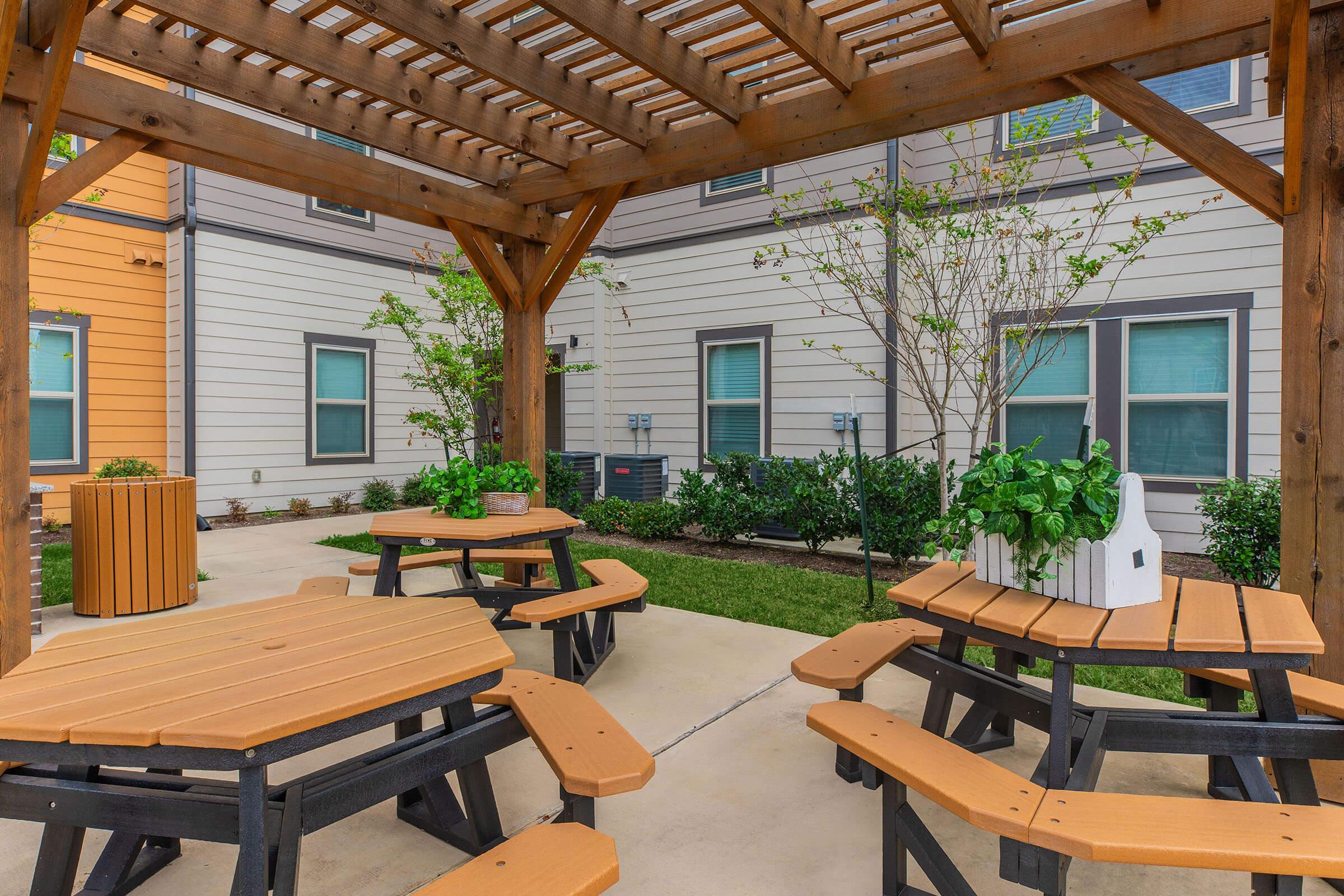 A cozy outdoor seating area with wooden picnic tables under a pergola. Surrounding greenery and buildings in the background create a welcoming atmosphere. A decorative planter is positioned on one of the tables, enhancing the space's charm.