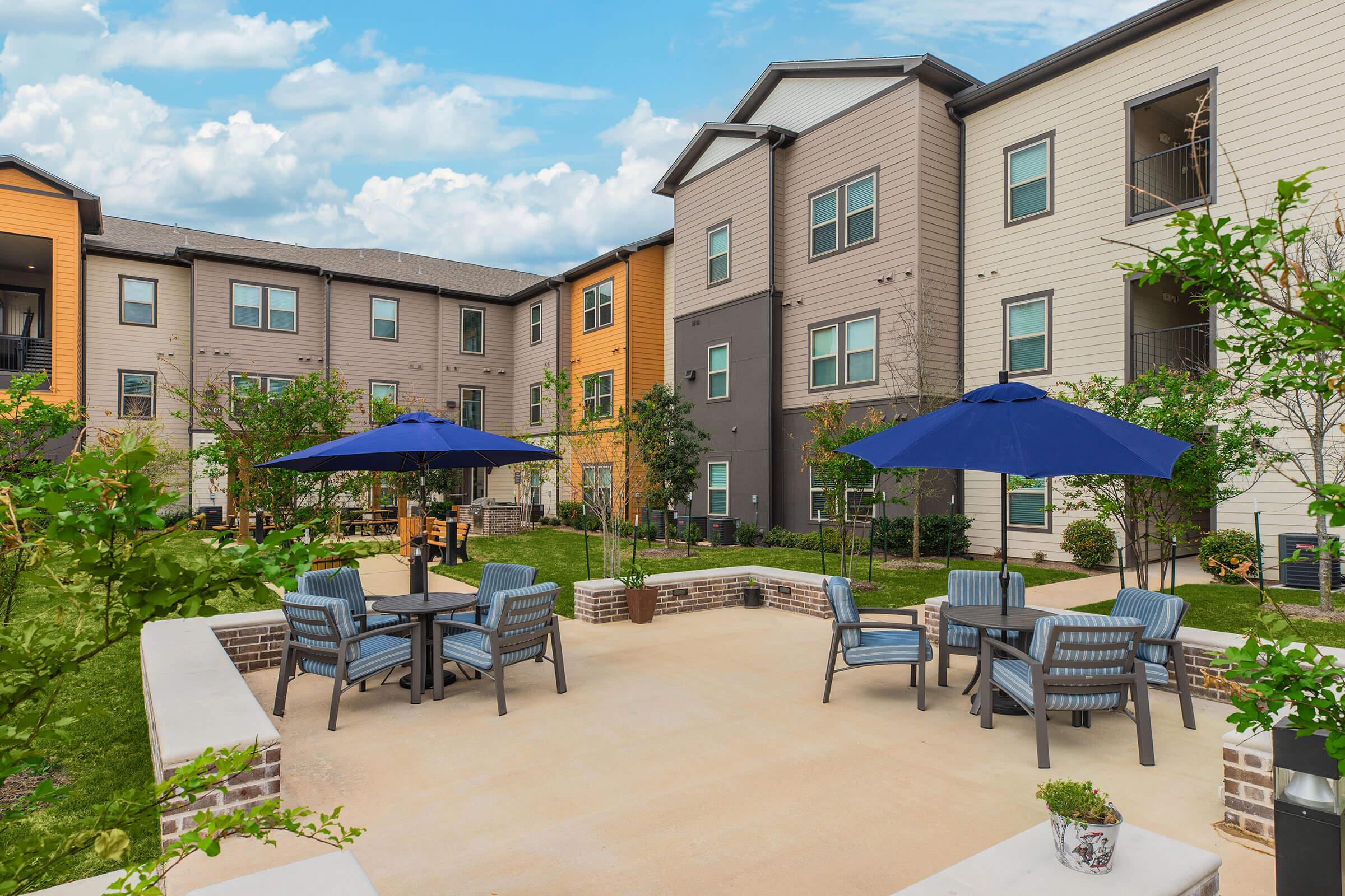 A landscaped courtyard with several tables and chairs under blue umbrellas. Surrounding buildings feature a mix of colors and styles. The area is well-maintained, with trees and greenery adding to the inviting atmosphere. The sky is partly cloudy, enhancing the outdoor setting.