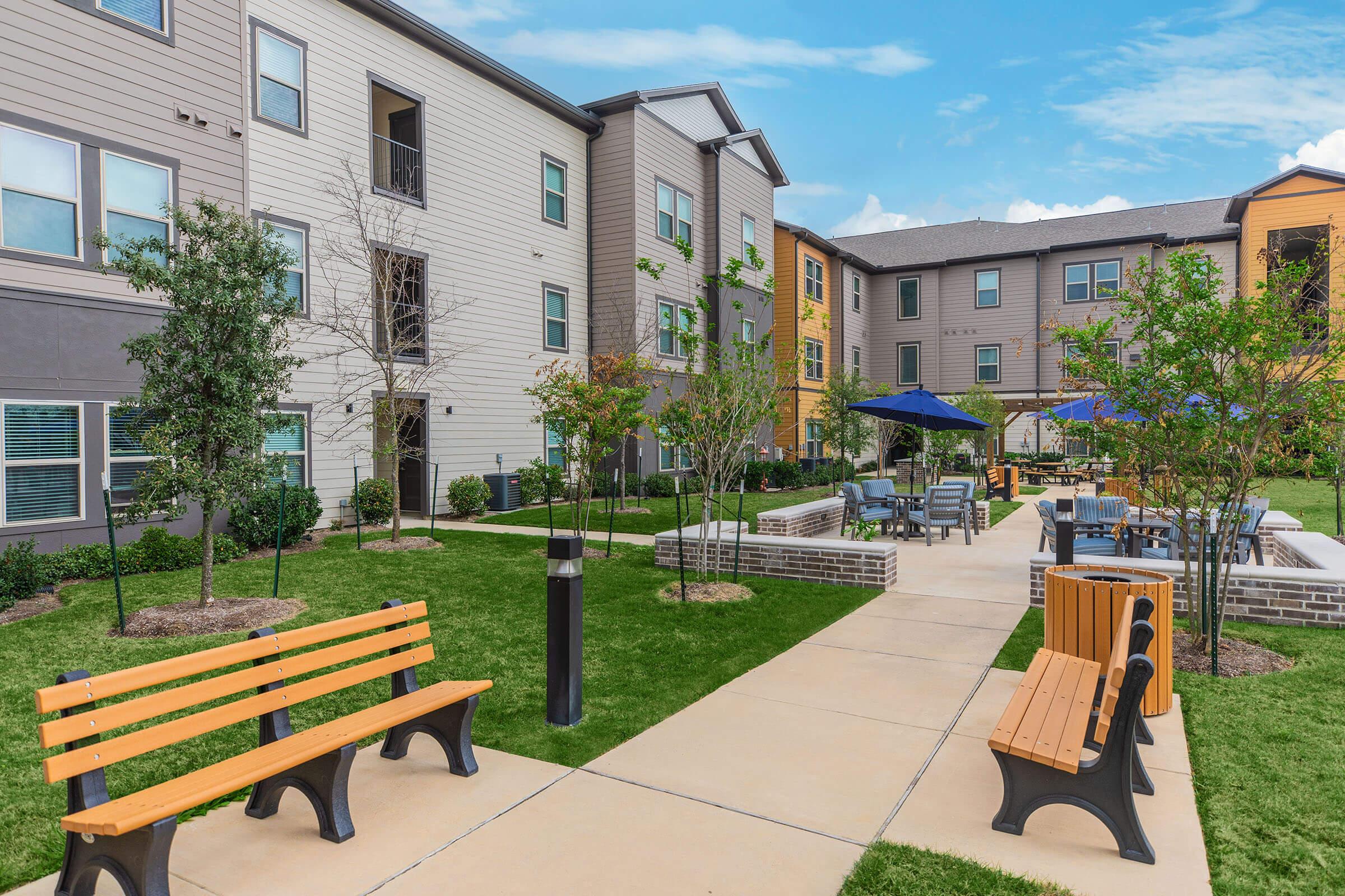 A landscaped courtyard of a modern apartment complex featuring several trees, benches, and tables with umbrellas. There are well-maintained pathways, green grass, and brick seating areas, creating a welcoming outdoor space for residents. Blue sky with scattered clouds serves as a backdrop.