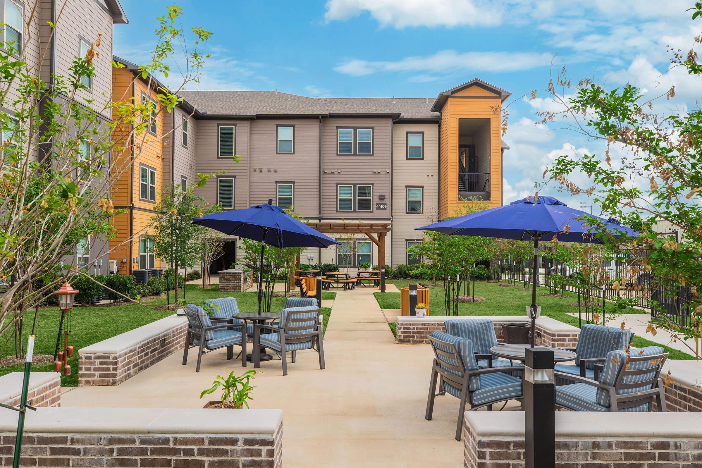 A landscaped courtyard featuring blue umbrellas over seating areas with chairs and tables. Surrounding greenery and trees add a natural touch, complemented by modern apartment buildings in the background. The sky is partly cloudy, creating a bright and inviting atmosphere.