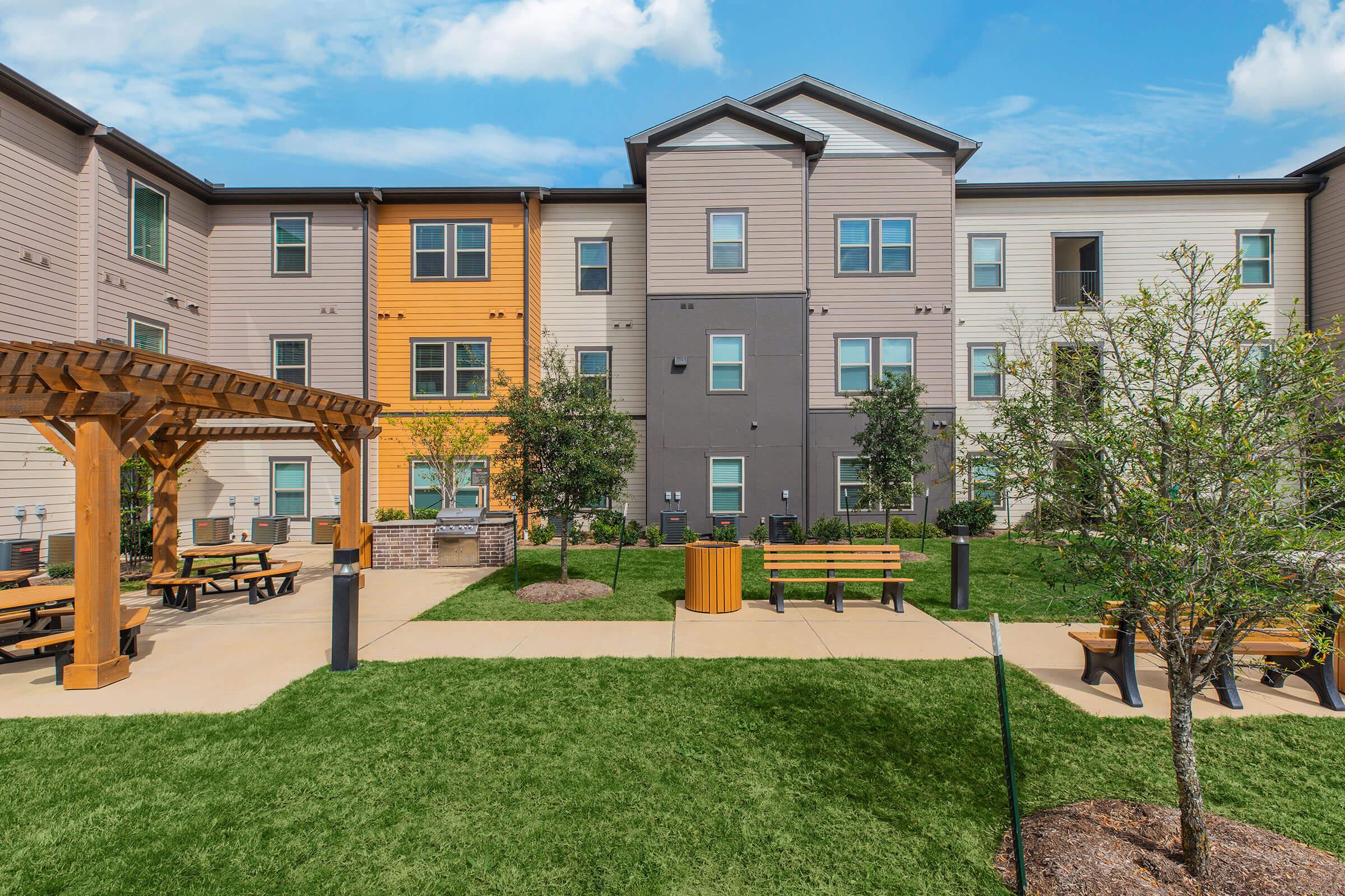 A modern apartment complex featuring a mix of beige, gray, and orange exteriors. The scene includes a landscaped courtyard with benches, a pergola, and trees. The sky is bright with clouds, emphasizing a welcoming outdoor space for residents.