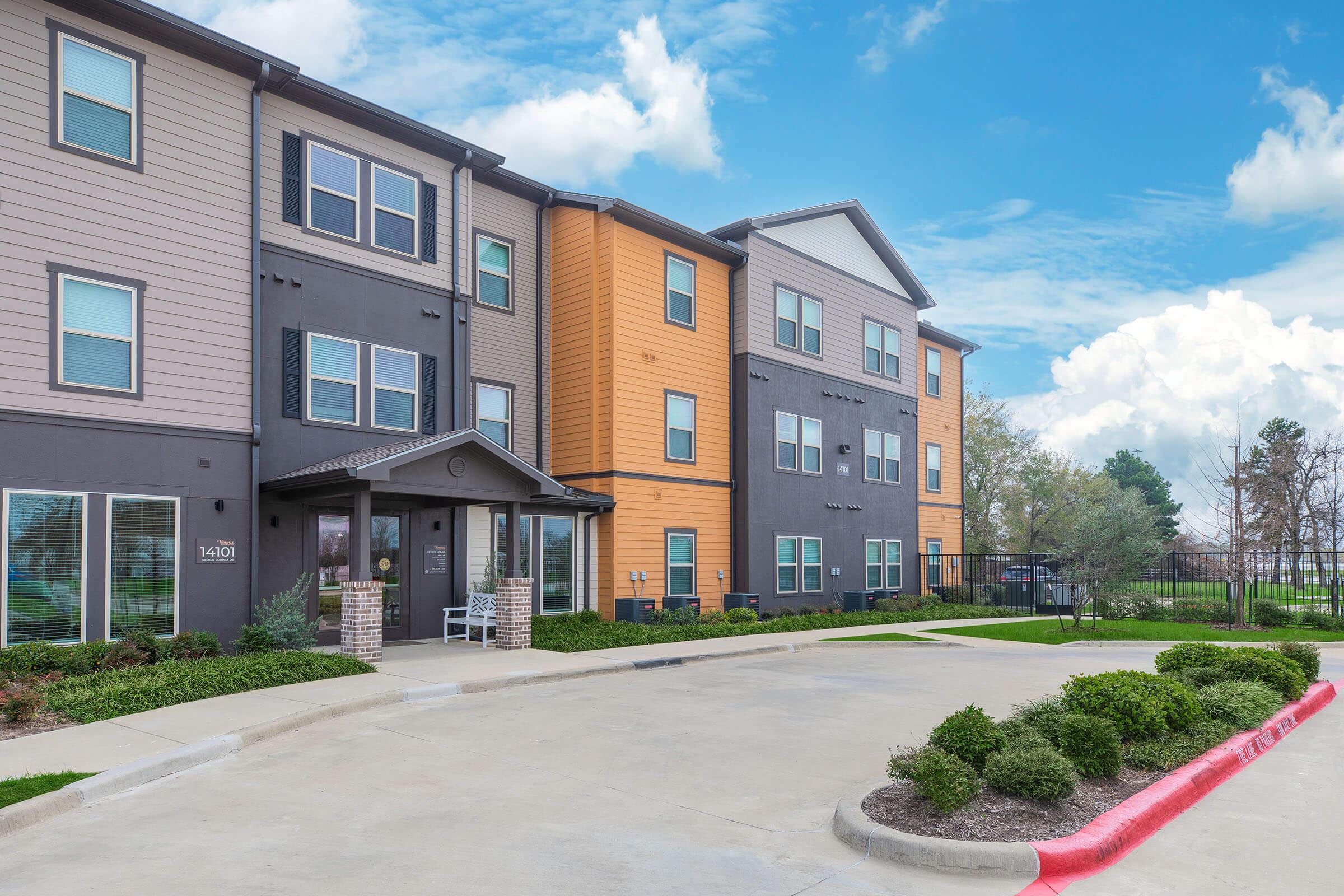 Modern apartment building with a mix of brown and orange siding, featuring multiple windows and an entrance awning. A paved parking area is visible in front, with well-maintained shrubs and landscaping. The sky is partly cloudy, adding a bright atmosphere to the scene.
