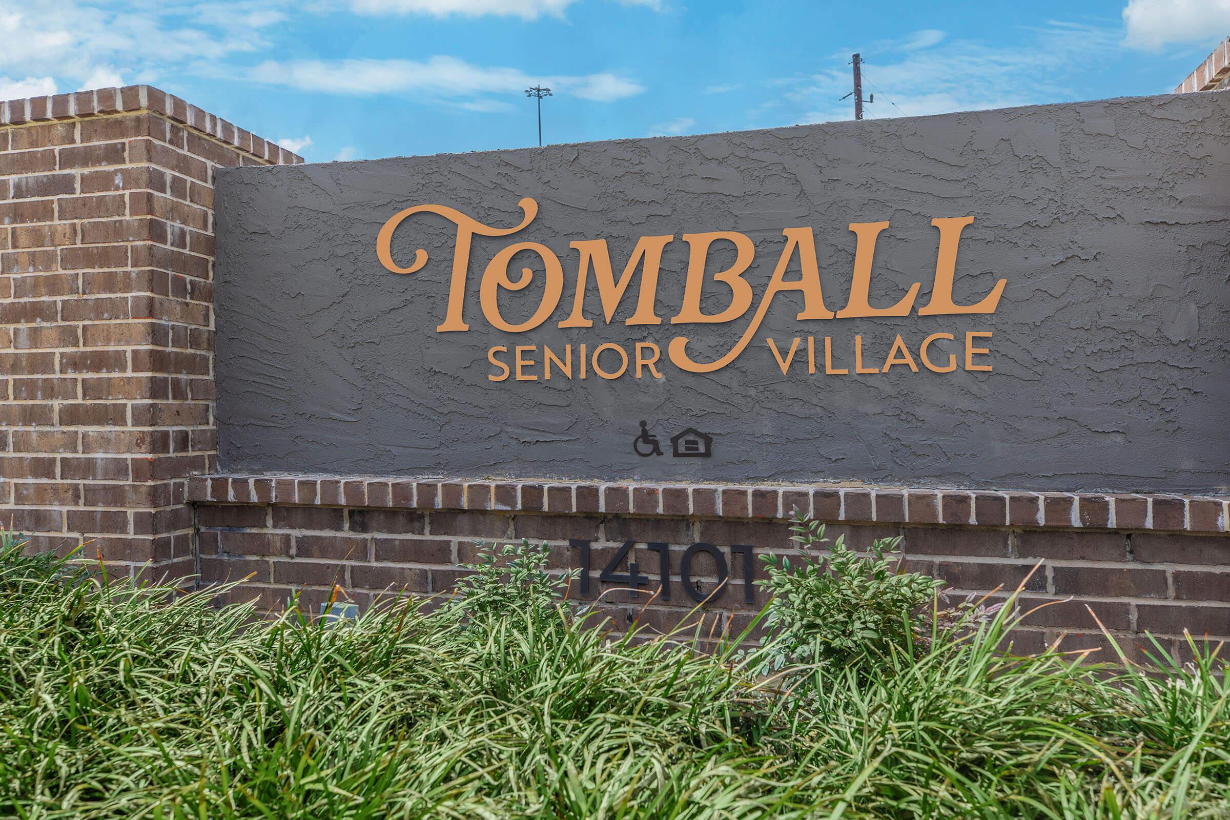 Sign for Tomball Senior Village, featuring the name in large, decorative letters. The sign is mounted on a brick structure, surrounded by green grass and plants, with a clear blue sky in the background. The address "1491" is displayed below the name.