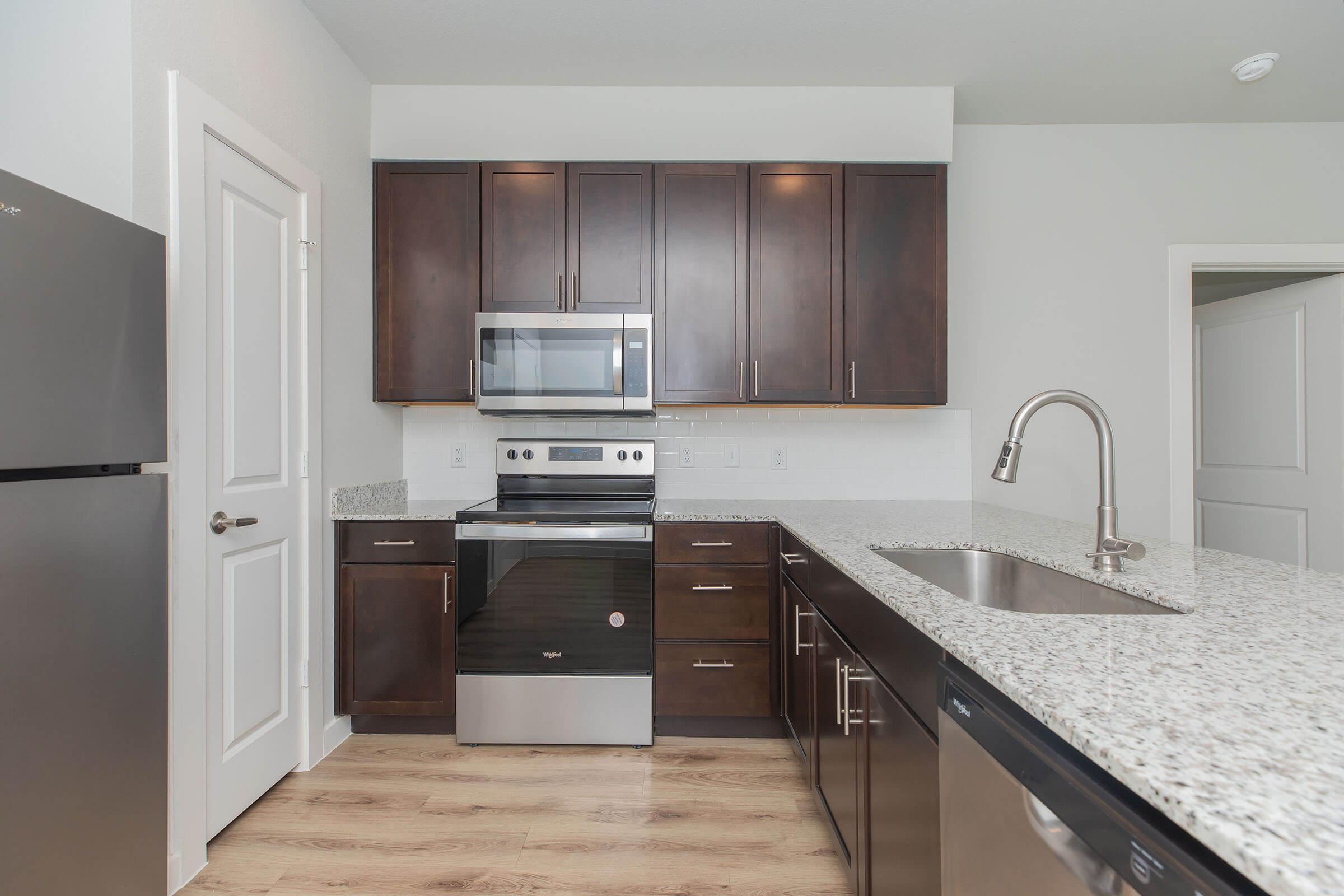 Modern kitchen featuring dark wooden cabinetry, a stainless steel microwave, an electric stove, and a sleek refrigerator. The countertop is made of light-colored granite, and there is a stainless steel sink with a faucet. The walls are painted light, creating a bright and inviting atmosphere.