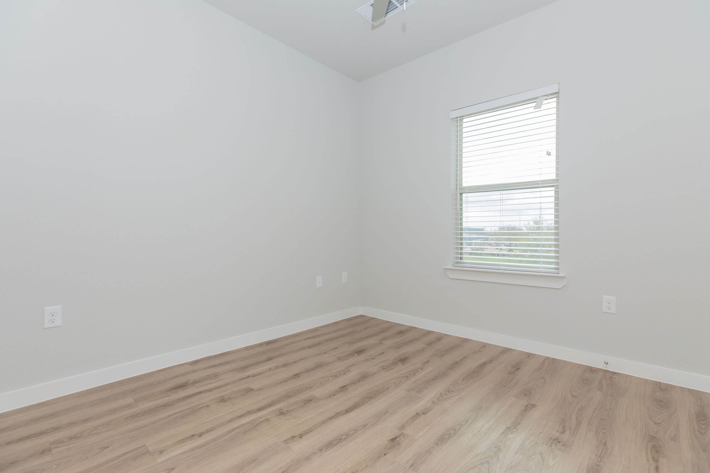 A vacant room featuring light-colored walls and wooden flooring. A window with blinds allows natural light to enter, creating a bright atmosphere. The space is minimalist and uncluttered, emphasizing its potential for various uses such as a bedroom or office.