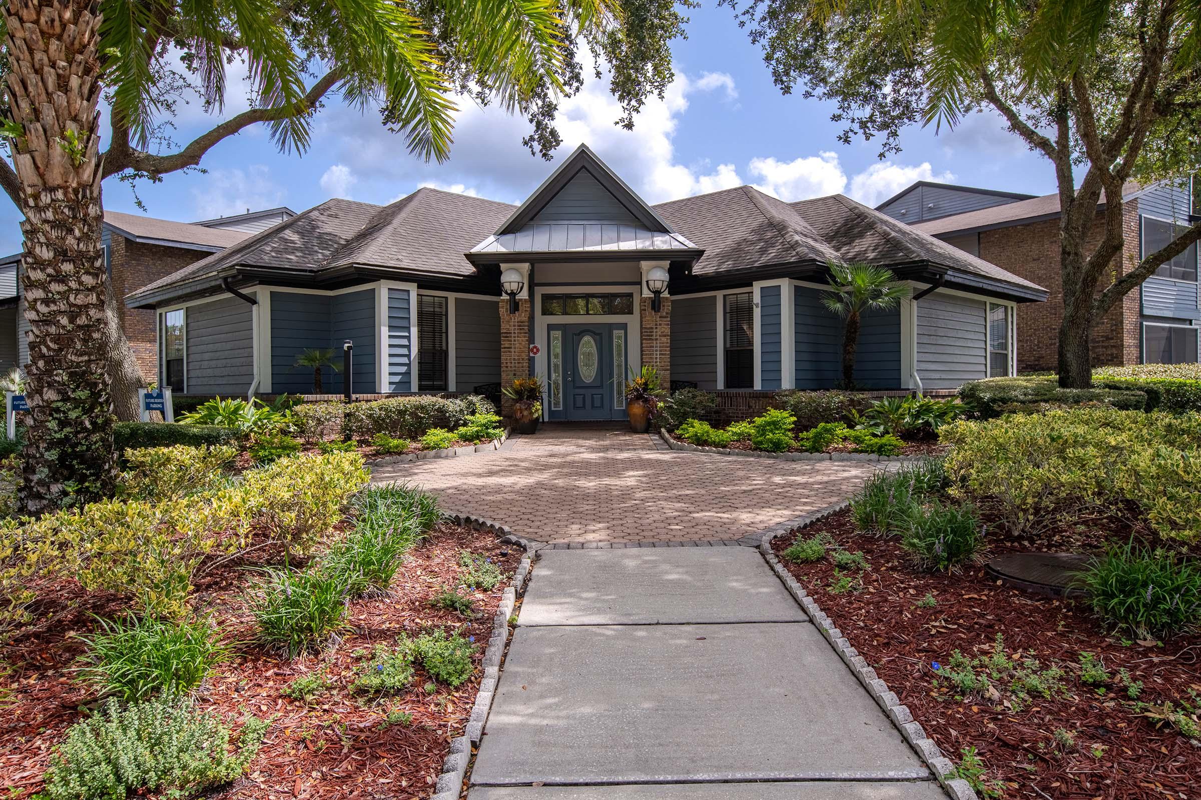 A modern blue and gray house with a peaked roof and a welcoming front porch. The landscaped yard features tropical plants, palm trees, and a stone pathway leading to the entrance. Bright blue sky with some fluffy clouds is visible in the background.