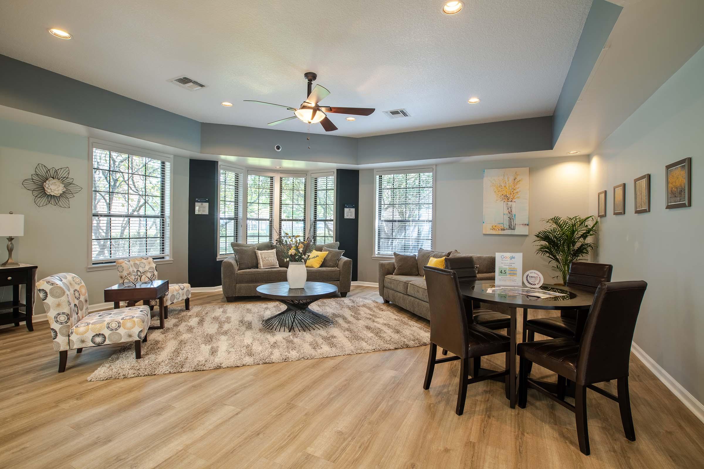 A cozy living room featuring a comfortable gray sofa with decorative pillows, two accent chairs, a round dining table with dark chairs, and a light rug. Large windows allow natural light, and the space is decorated with artwork and plants, creating a warm and inviting atmosphere.