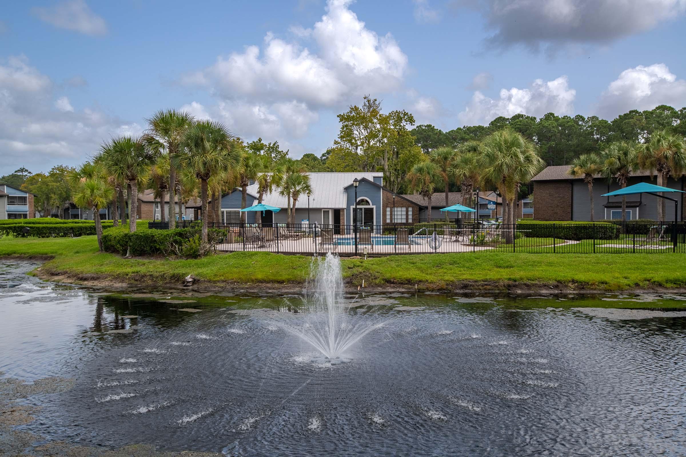 A serene residential area featuring a pond with a fountain at the center. The background showcases well-maintained lawns, palm trees, and apartment buildings. Clear blue skies with scattered clouds add to the tranquil atmosphere, while lounge areas with umbrellas are visible near the pool.