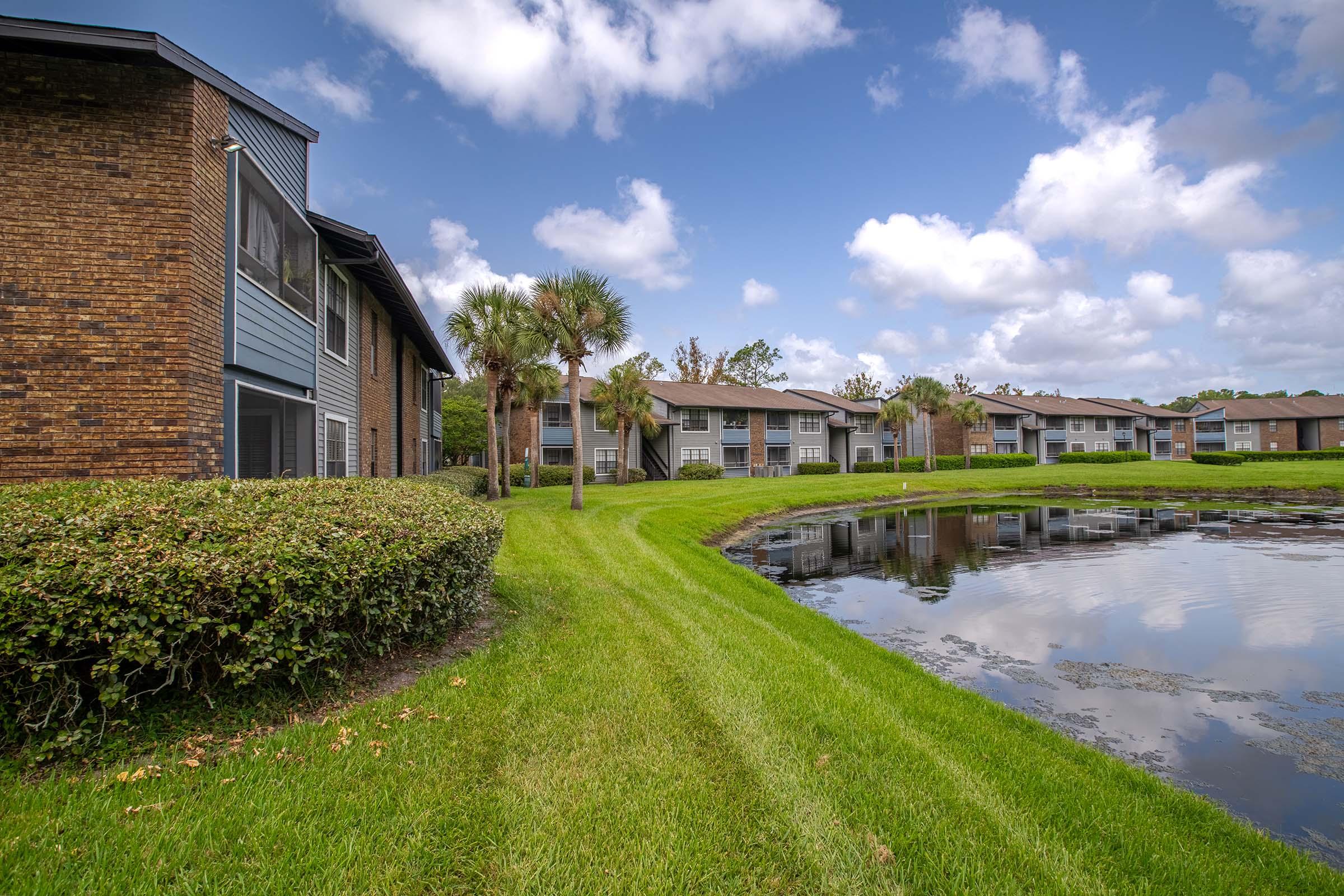 A scenic view of a residential area featuring multi-story buildings alongside a small pond. Lush green grass and bushes line the pathway next to the water, with palm trees visible among the landscape. The sky is partly cloudy, creating a serene atmosphere.