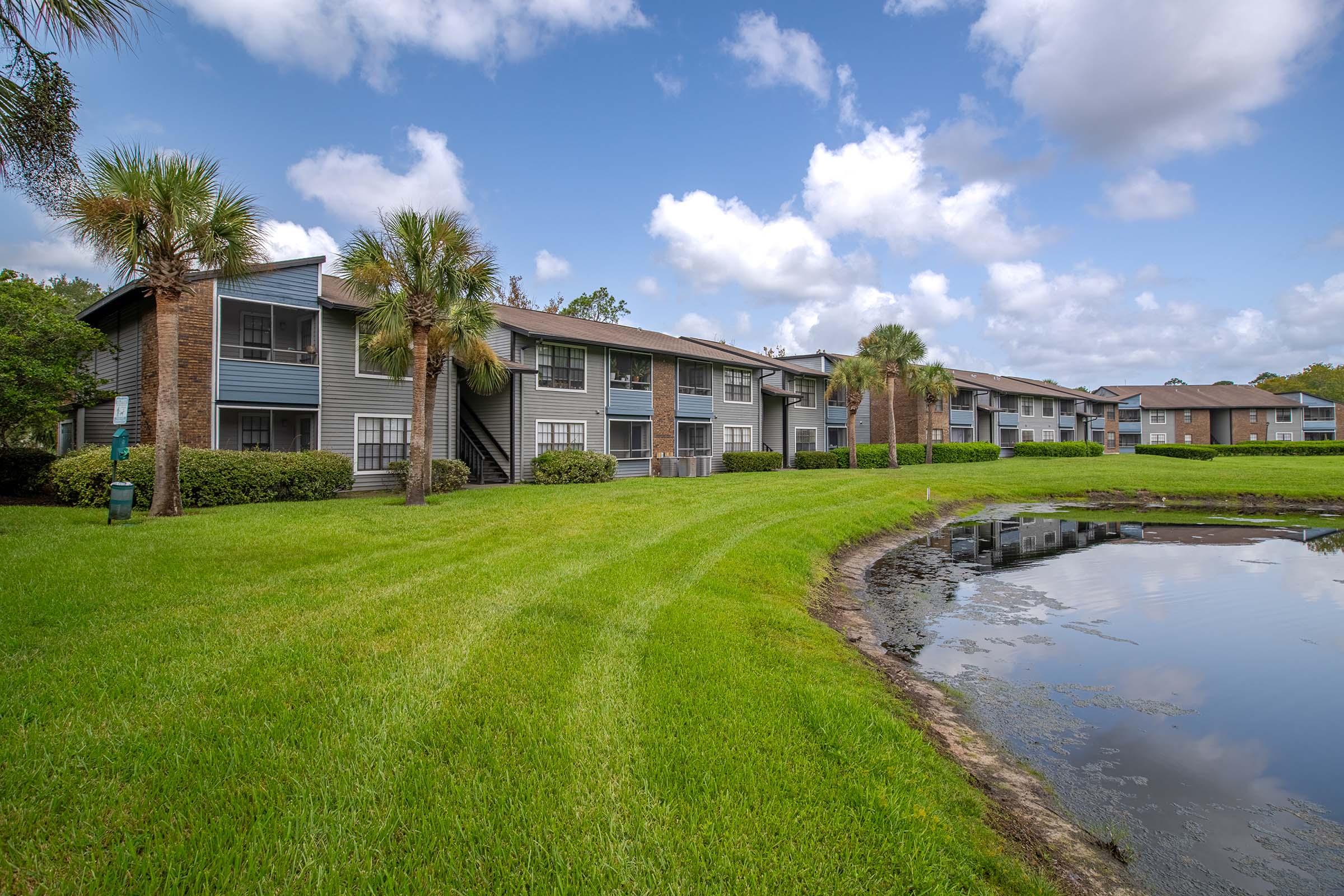A grassy area with palm trees and a calm pond in front of several apartment buildings. The sky is partly cloudy, and the landscape is well-maintained, showcasing a serene residential setting.