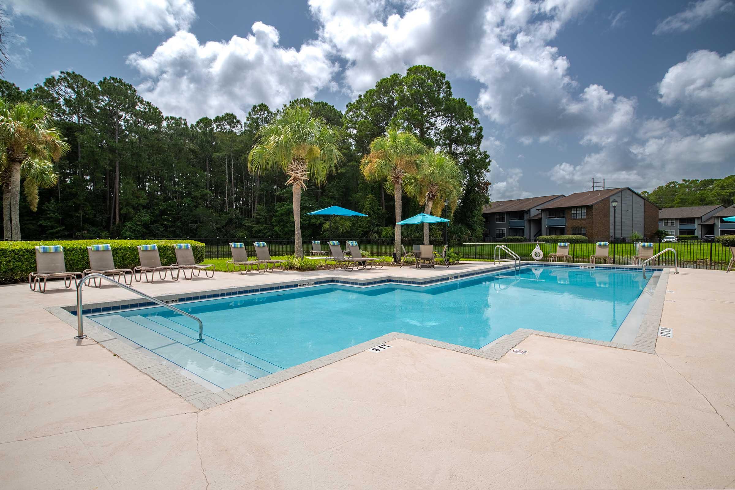 A sparkling blue swimming pool surrounded by lounge chairs and palm trees, set against a backdrop of green foliage and a partly cloudy sky. Two shaded umbrellas are positioned near the pool, creating a relaxing atmosphere in a residential area.