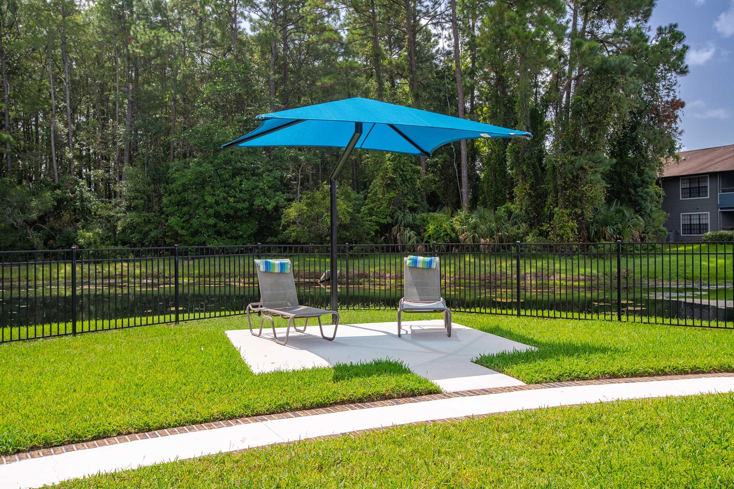 Two lounge chairs with cushions positioned under a bright blue umbrella, set on a concrete pad surrounded by lush green grass. In the background, a wooded area is visible, alongside a fenced-in seating area near a body of water. The sky is clear with a few clouds.