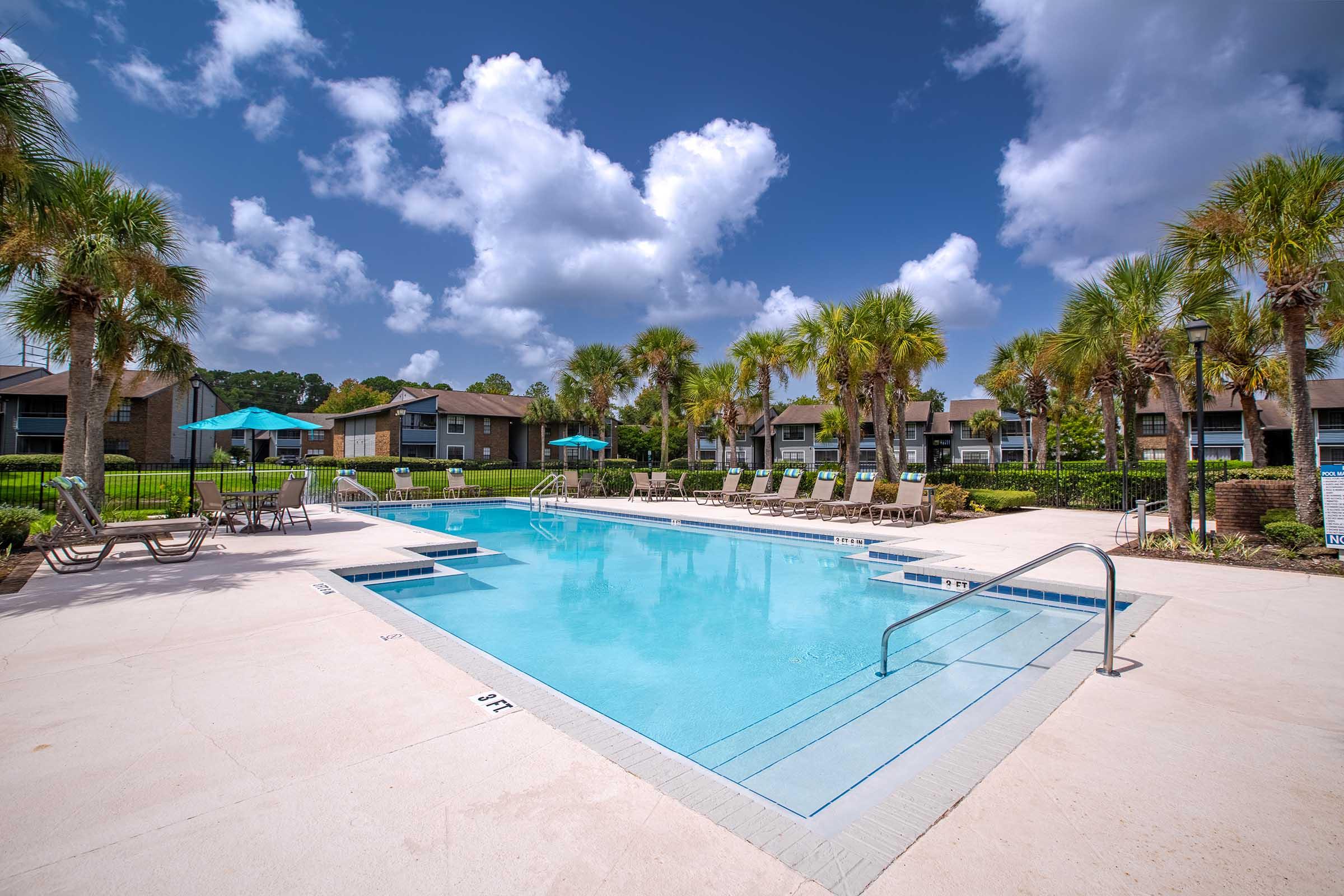 A clear swimming pool surrounded by lounge chairs and umbrellas, with palm trees lining the area. The sky is bright blue with fluffy white clouds, and there are residential buildings in the background. The scene conveys a relaxing outdoor environment.