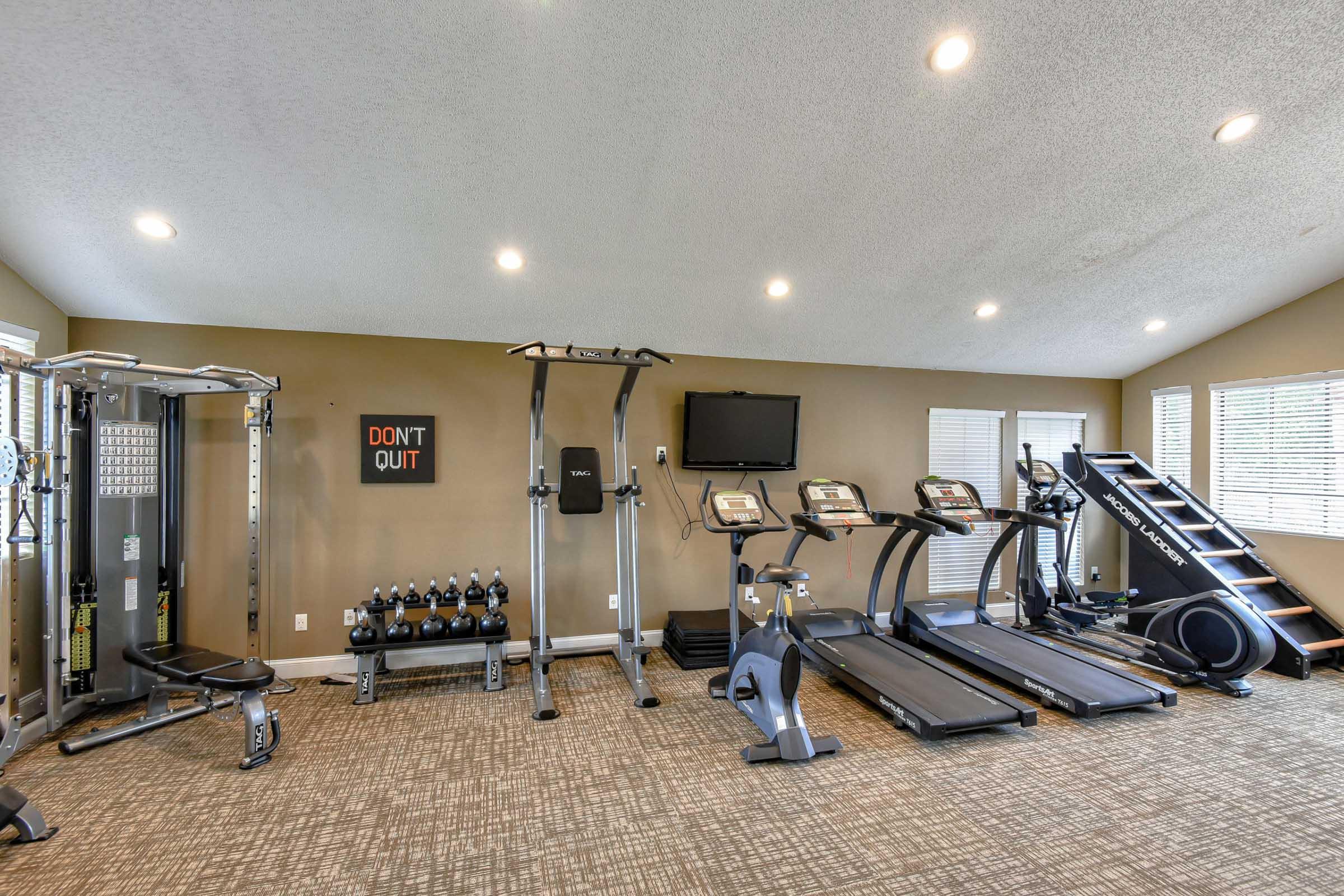A well-equipped fitness room featuring various exercise machines, including treadmills, an elliptical trainer, a cable machine, and free weights. The space is brightly lit with large windows and motivational wall art that reads "DON'T QUIT." The flooring is carpeted for comfort.