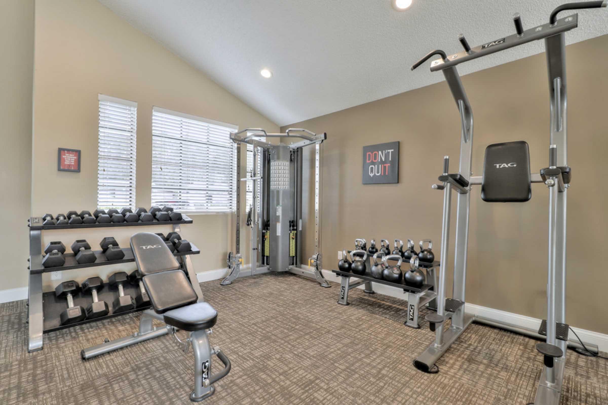 A modern fitness room featuring various exercise equipment. The room includes a weight bench, dumbbell rack with weights, a multi-station gym machine, and a motivational poster that says "DON'T QUIT." Natural light streams in through several windows, enhancing the workout space ambiance.