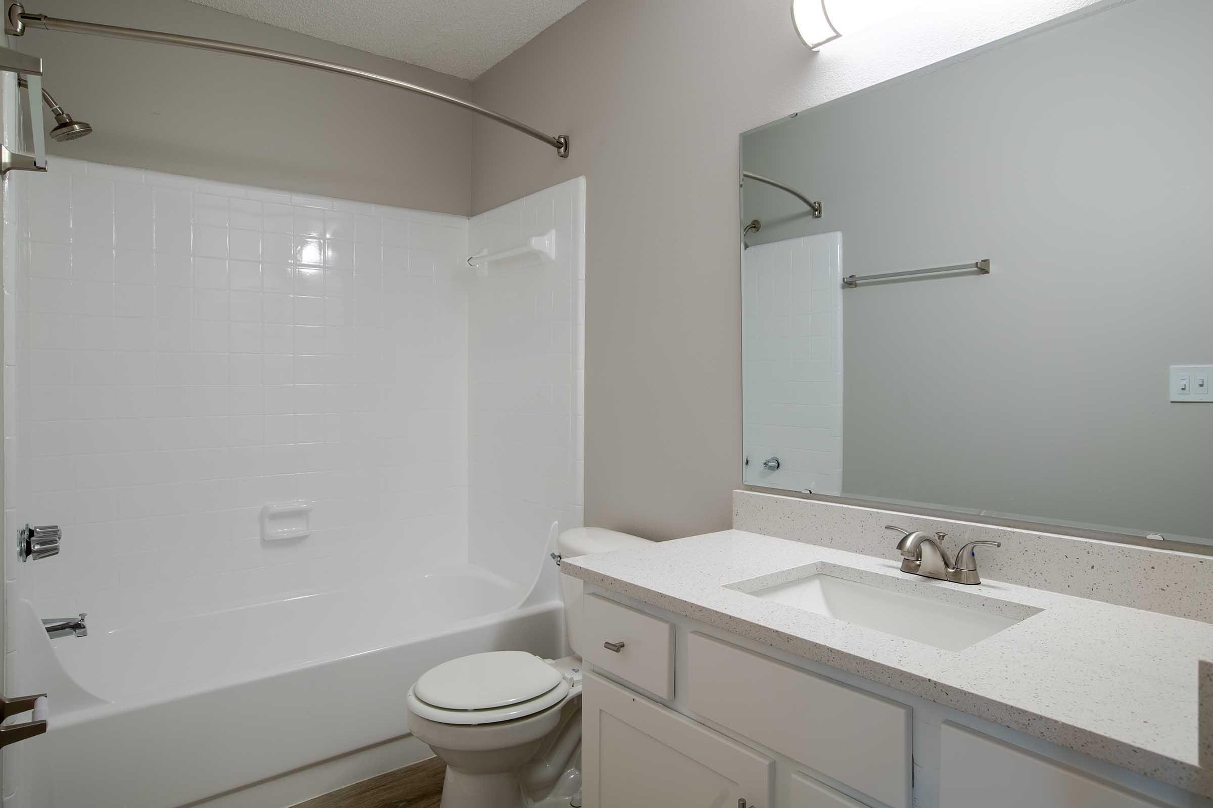 A clean, modern bathroom featuring a white bathtub with a shower, a white vanity with a sink, and a large mirror above it. The walls are painted in a soft gray, and the floor has a light wood finish. A towel bar is mounted on the wall beside the mirror.