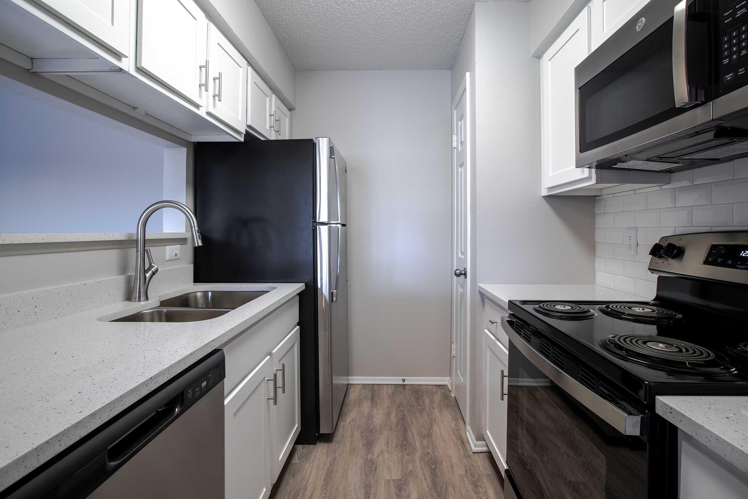 A modern kitchen featuring white cabinetry, stainless steel appliances including a refrigerator, microwave, and stove, a double sink, and dark countertops. The space is designed with a light color scheme and has a wood-like floor, creating a bright and inviting atmosphere.