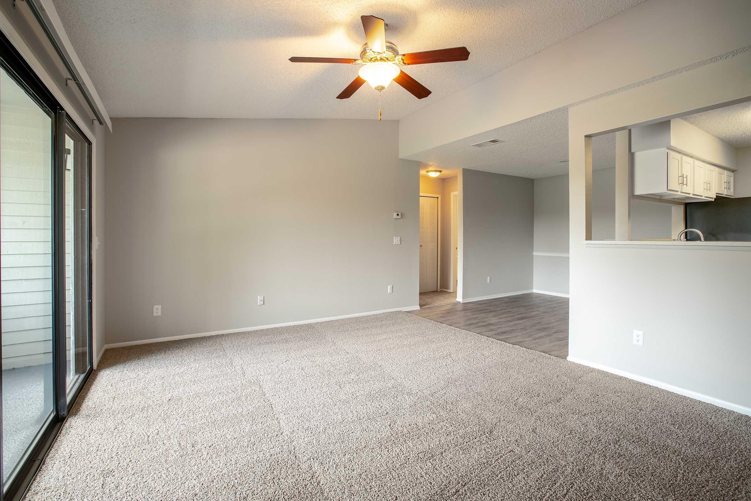 Bright and spacious living area with ceiling fan, light-colored carpet, and neutral walls. Features large sliding glass doors leading to a balcony. Open layout connecting to the kitchen, with a doorway visible in the background. Smooth flooring transitions to the kitchen area.