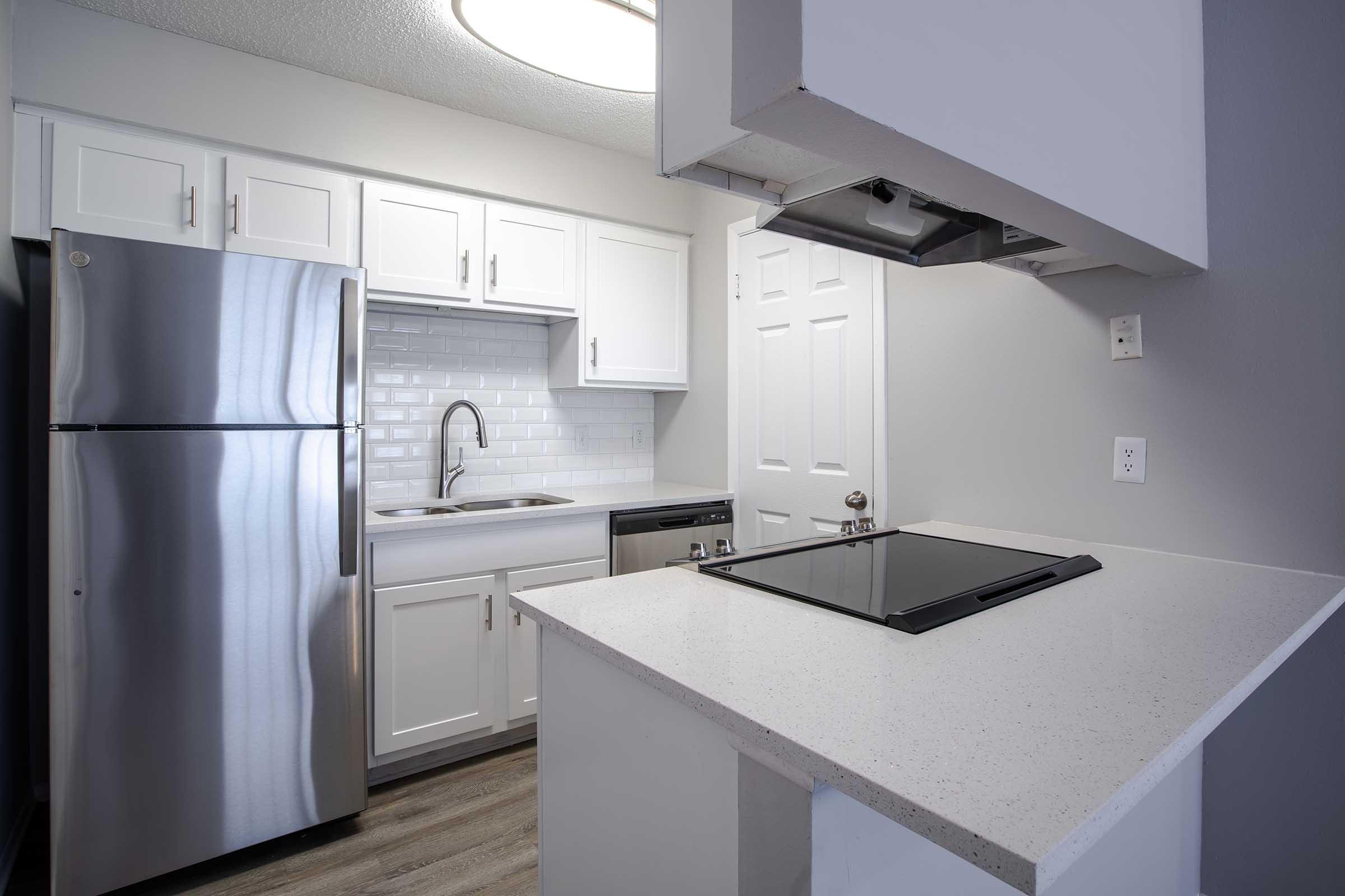 Modern kitchen featuring stainless steel appliances, including a refrigerator and dishwasher. The space has white cabinets, a sink with a sleek faucet, and a cooktop on a white countertop. The backsplash consists of white subway tiles, and soft overhead lighting enhances the bright, clean atmosphere.