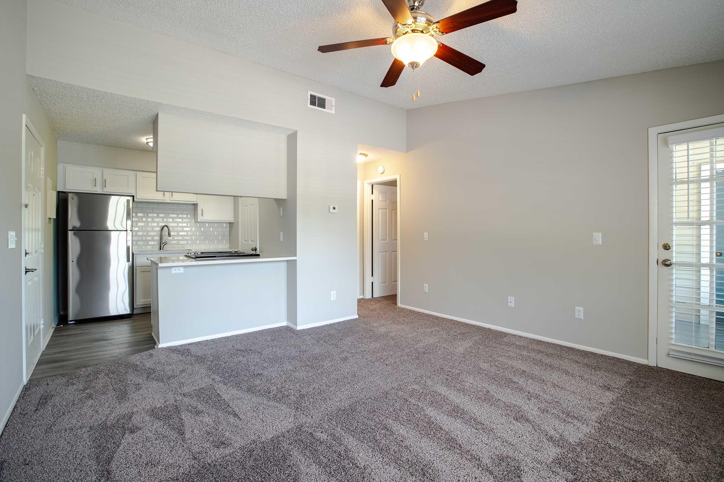 Spacious living area with carpeted flooring, a ceiling fan, and a view of the kitchen. The kitchen features modern appliances and white cabinetry. A doorway leads to a private exterior space, and there are light-colored walls enhancing the bright atmosphere.
