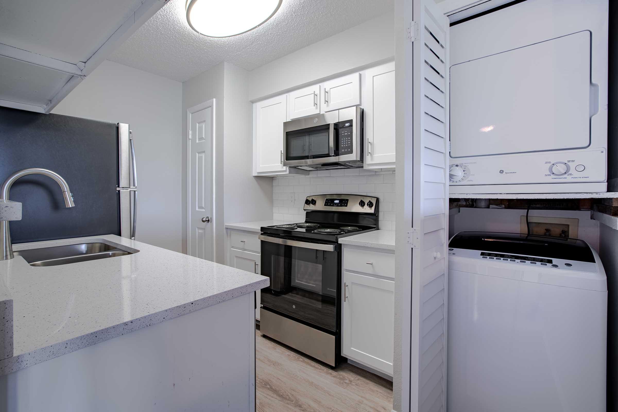 A modern kitchen featuring white cabinets, a stainless steel refrigerator, an oven, and a microwave. A countertop with a sink is visible in the foreground, while a stacked washer and dryer are situated beside the kitchen area. The space has neutral-colored walls and a bright overhead light.
