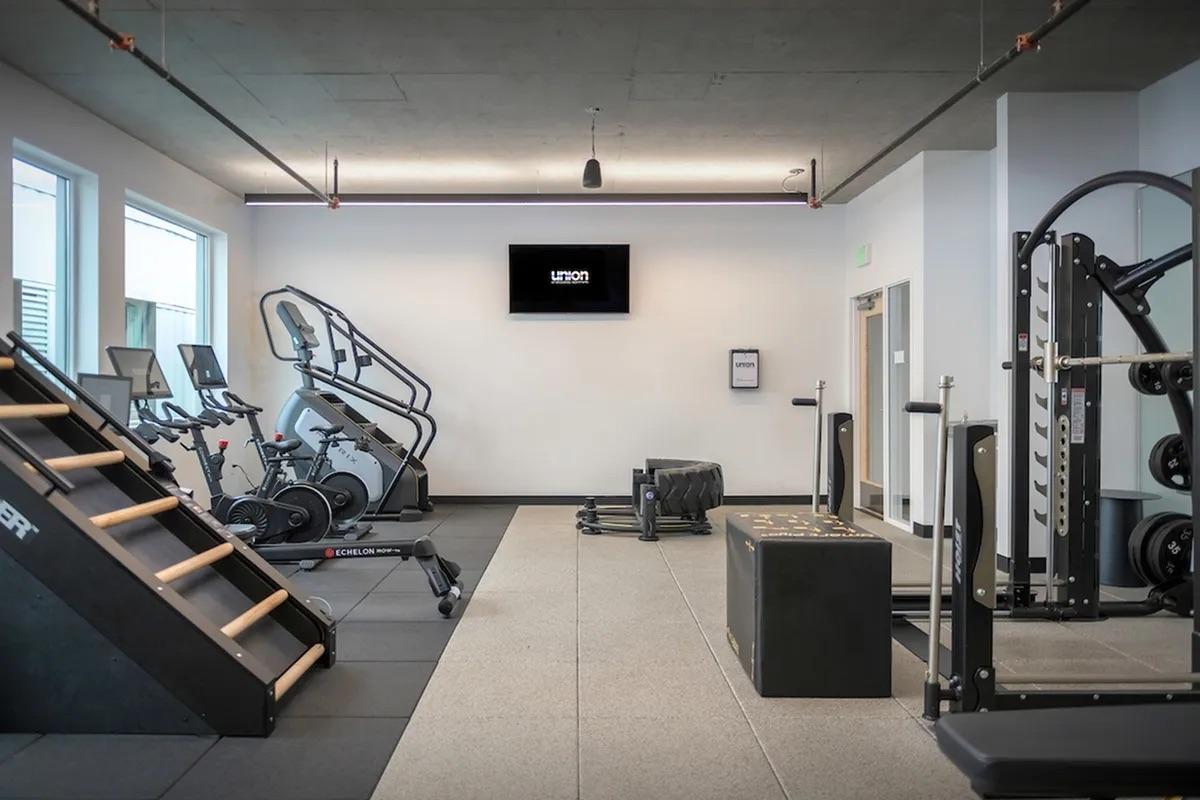 A modern gym interior featuring various exercise equipment. There are stair climbers, stationary bikes, a weight machine, and a functional training area with a sled and a box for workouts. The walls are painted light colors, and a flat-screen TV hangs on one wall. The flooring consists of dark rubber tiles.