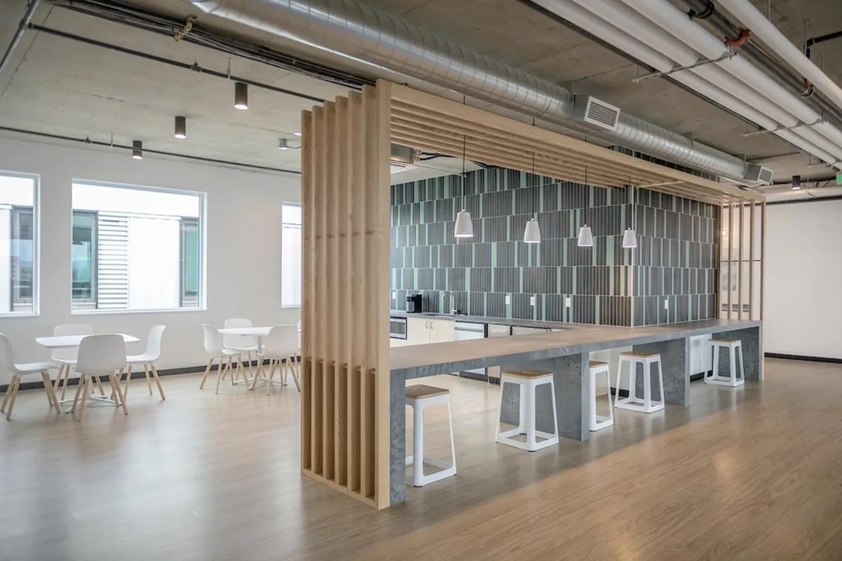 A modern, minimalist kitchen area featuring a long countertop with a wood and concrete design. The space includes white chairs and tables, pendant lighting, and a sleek tile backsplash. Large windows let in natural light, enhancing the contemporary feel of the office environment.
