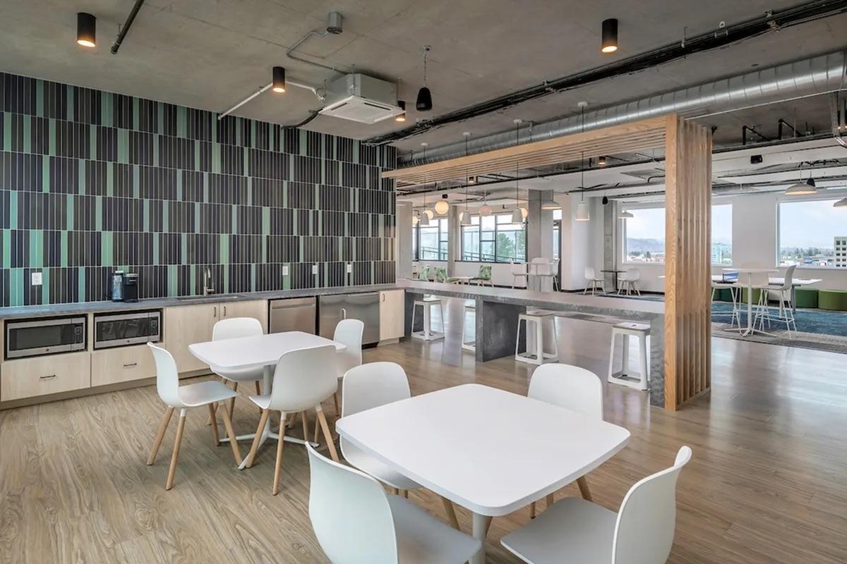 Modern office break room featuring a sleek design with light wood floors, white tables and chairs, and a kitchen area with cabinets and appliances. The walls are adorned with a patterned design in shades of green and black, while large windows allow natural light to fill the space.