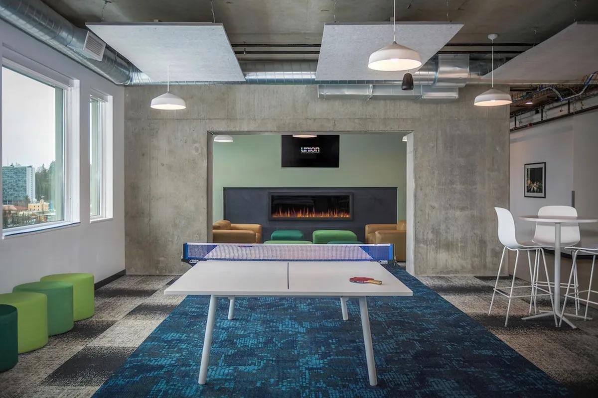 A modern office space featuring a ping-pong table in the center, surrounded by contemporary furniture. In the background, a cozy lounge area with two brown sofas, a fireplace, and a large TV mounted on a concrete wall. The floors are a mix of carpet and tiles, with green stools and a white table nearby.