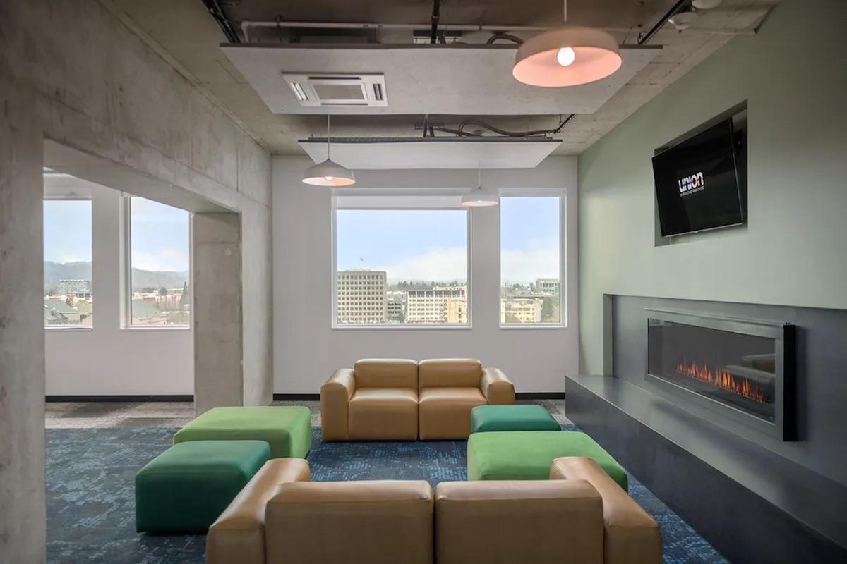 A modern lounge area featuring a tan leather sofa, green poufs, large windows with a city view, and a wall-mounted TV above an electric fireplace. The room has a contemporary design with concrete walls and a stylish carpet.