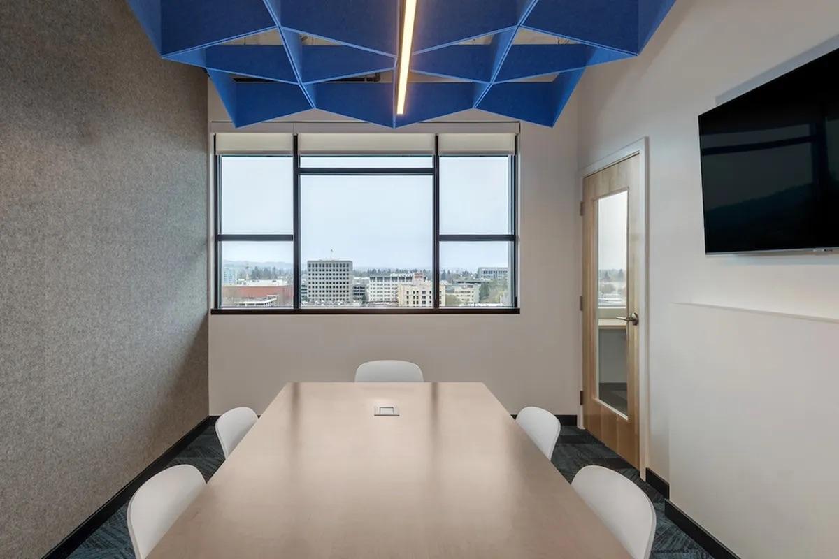 A modern conference room featuring a long wooden table surrounded by white chairs. The room has large windows showing a city skyline view, with a blue geometric ceiling design and a wall-mounted television. The overall ambiance is bright and professional.