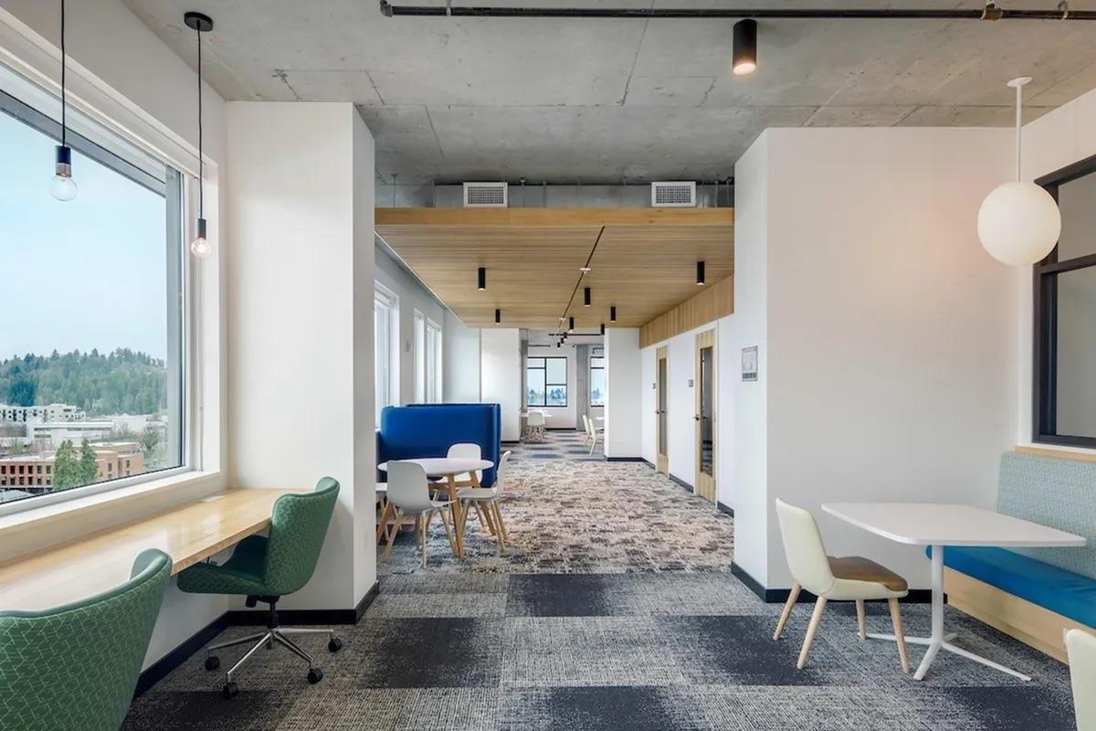 Modern office interior featuring a spacious, open layout. Includes a carpeted area with contrasting tile floors, large windows, and wooden ceiling accents. Furniture includes a round wooden table with chairs, a blue upholstered booth, and workstations against the wall, creating a collaborative workspace environment.