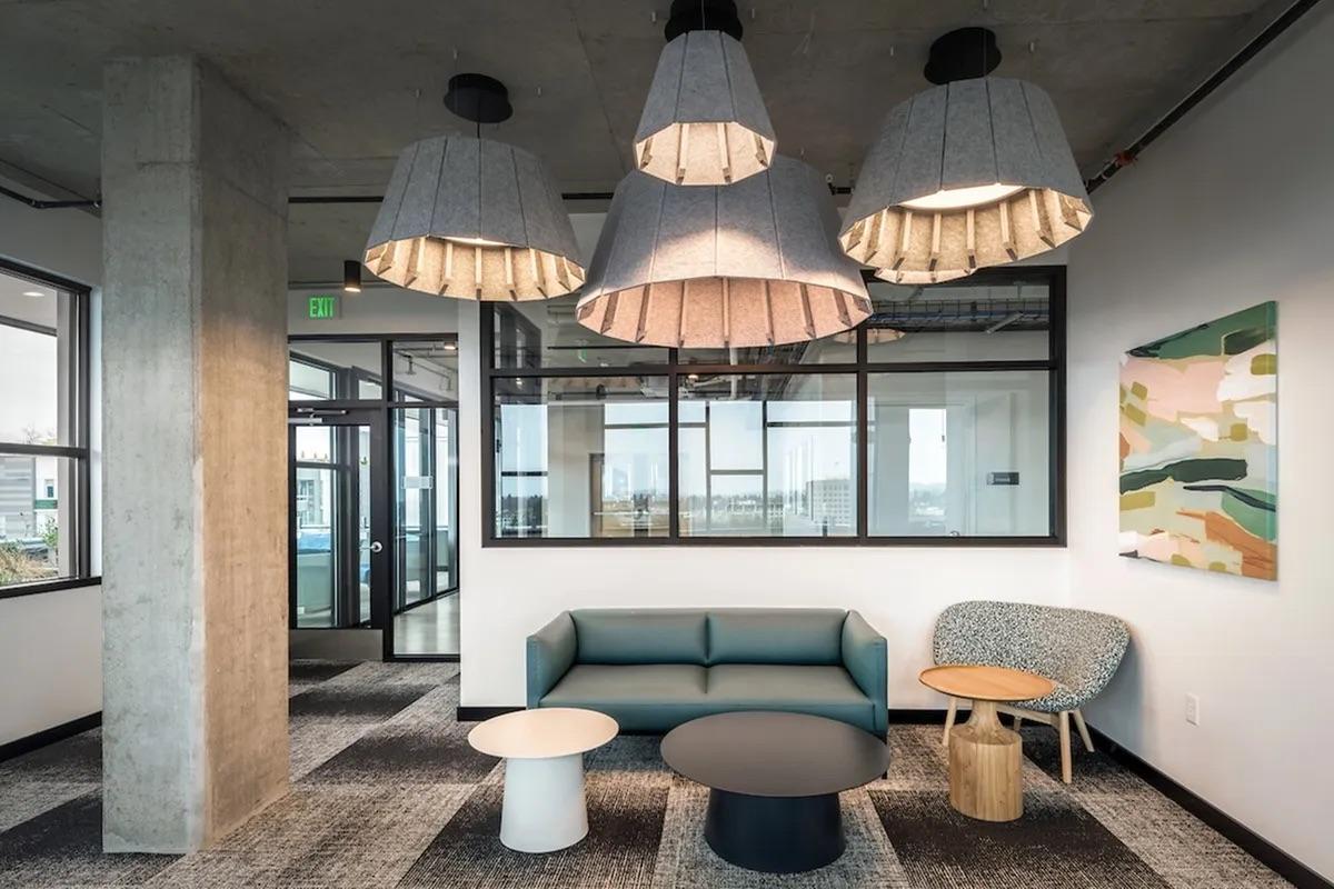 A modern office lounge featuring a blue sofa, round black coffee table, light wood side table, and patterned chair. The space is illuminated by large, hanging lamps with fabric shades. Large windows provide natural light, and a colorful abstract artwork adorns the wall. The flooring is a mix of carpet textures.