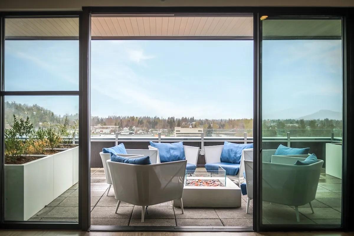 A modern outdoor patio with a seating arrangement featuring white chairs and a coffee table adorned with a decorative item. The view showcases a serene landscape with trees and distant mountains under a clear sky. Glass doors frame the scene, creating an inviting atmosphere for relaxation.