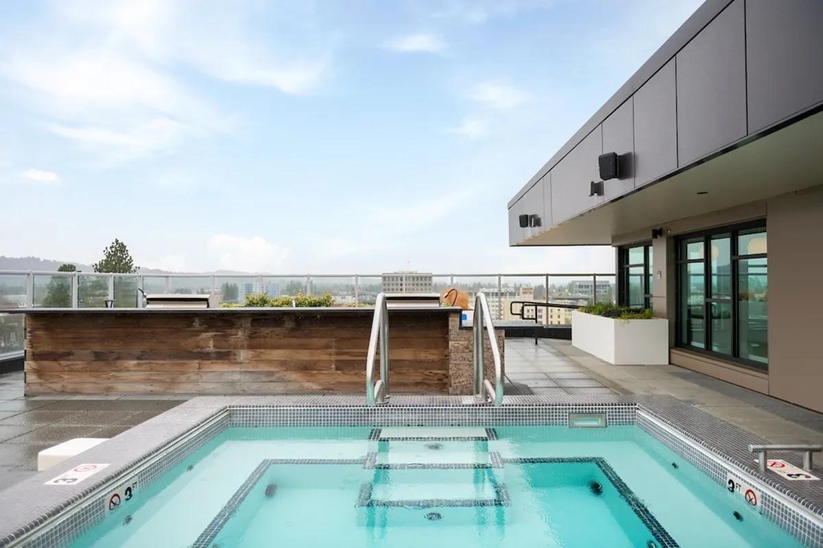 Rooftop area featuring a small spa pool with steps, surrounded by modern architectural elements. In the background, a panoramic view of the city skyline under a clear sky. The space appears well-designed for relaxation, with greenery and outdoor seating areas.