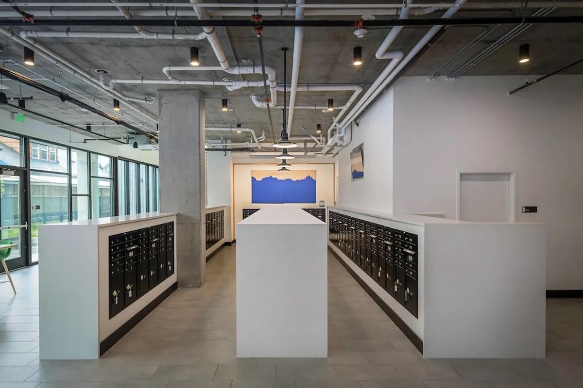 Modern indoor space featuring a row of black mailboxes along white walls. Concrete pillars and exposed pipes add an industrial feel. Large windows allow natural light, and a blue abstract artwork is displayed on the wall in the background. The floor is tiled in gray, creating a clean, minimalist aesthetic.