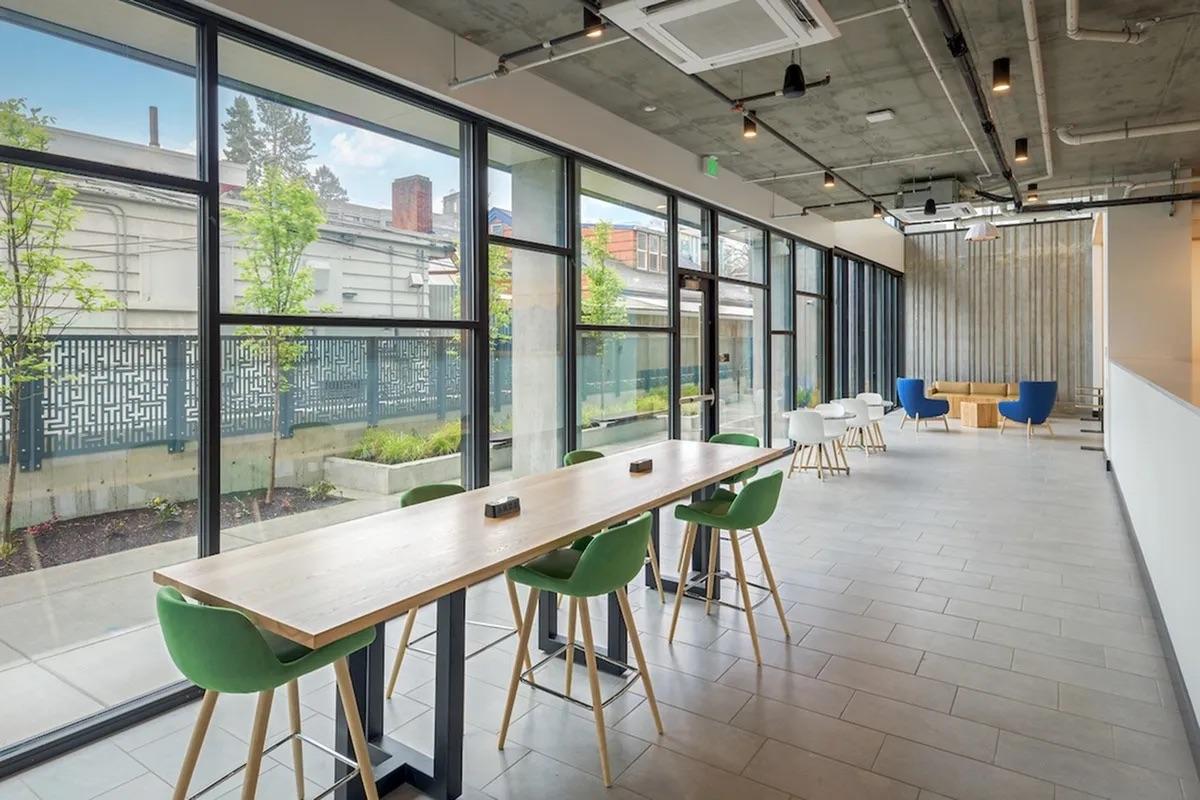 A modern, well-lit interior space featuring a long wooden table with green and white chairs, large windows allowing natural light, and a minimalist design. The area includes potted plants and a cozy seating arrangement, creating an inviting atmosphere for relaxation or work.