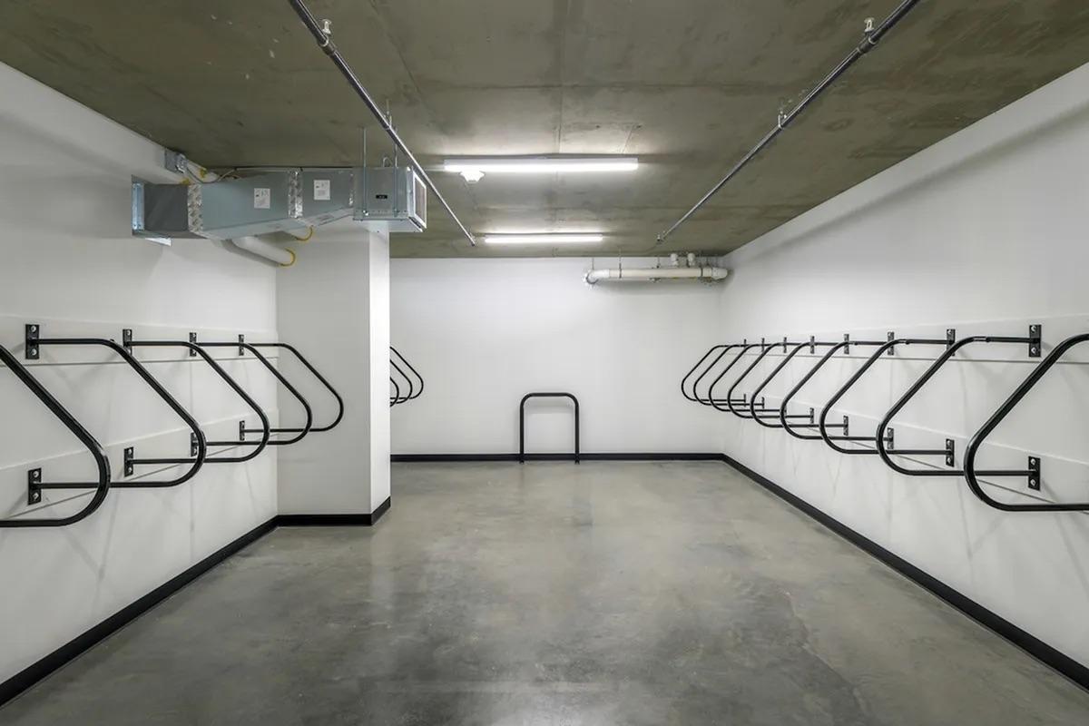 Empty bike storage room with metal bike racks mounted on the walls. The floor is concrete, and the walls are painted white. Ceiling lights illuminate the space, creating a bright and minimalist environment.