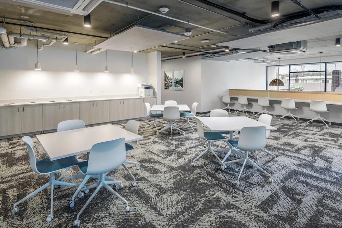 A modern office break room featuring several white tables and chairs arranged in a spacious layout. The floor has a patterned carpet, and there are cabinets along one wall. Soft lighting hangs from the ceiling, providing a bright atmosphere. A large window lets in natural light, and a water cooler is visible.