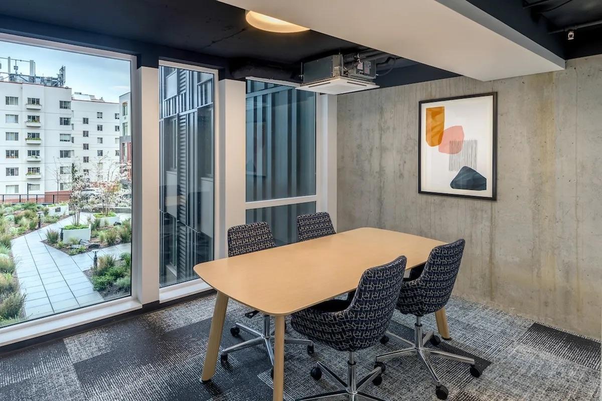 A modern conference room featuring a wooden table and four ergonomic chairs. Large windows provide natural light and a view of an outdoor garden area. Concrete walls and artwork add a contemporary touch to the space.